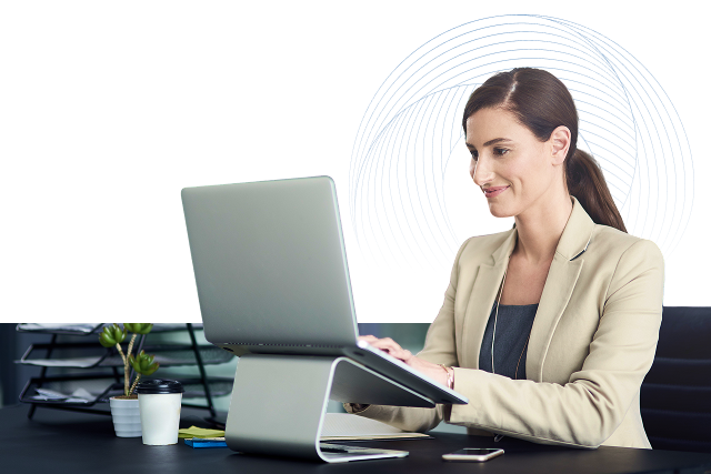 Frau sitzt lächelnd am Laptop an einem erhöhten Laptop-Ständer, im Hintergrund eine Pflanze