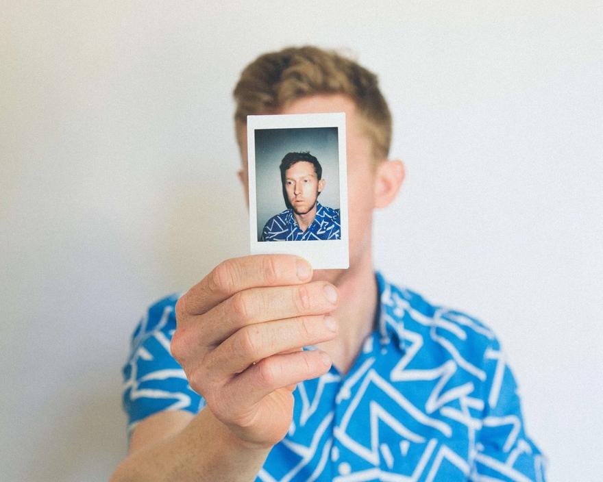 Man in a blue patterned shirt holds a portrait photo of himself in front of his face.