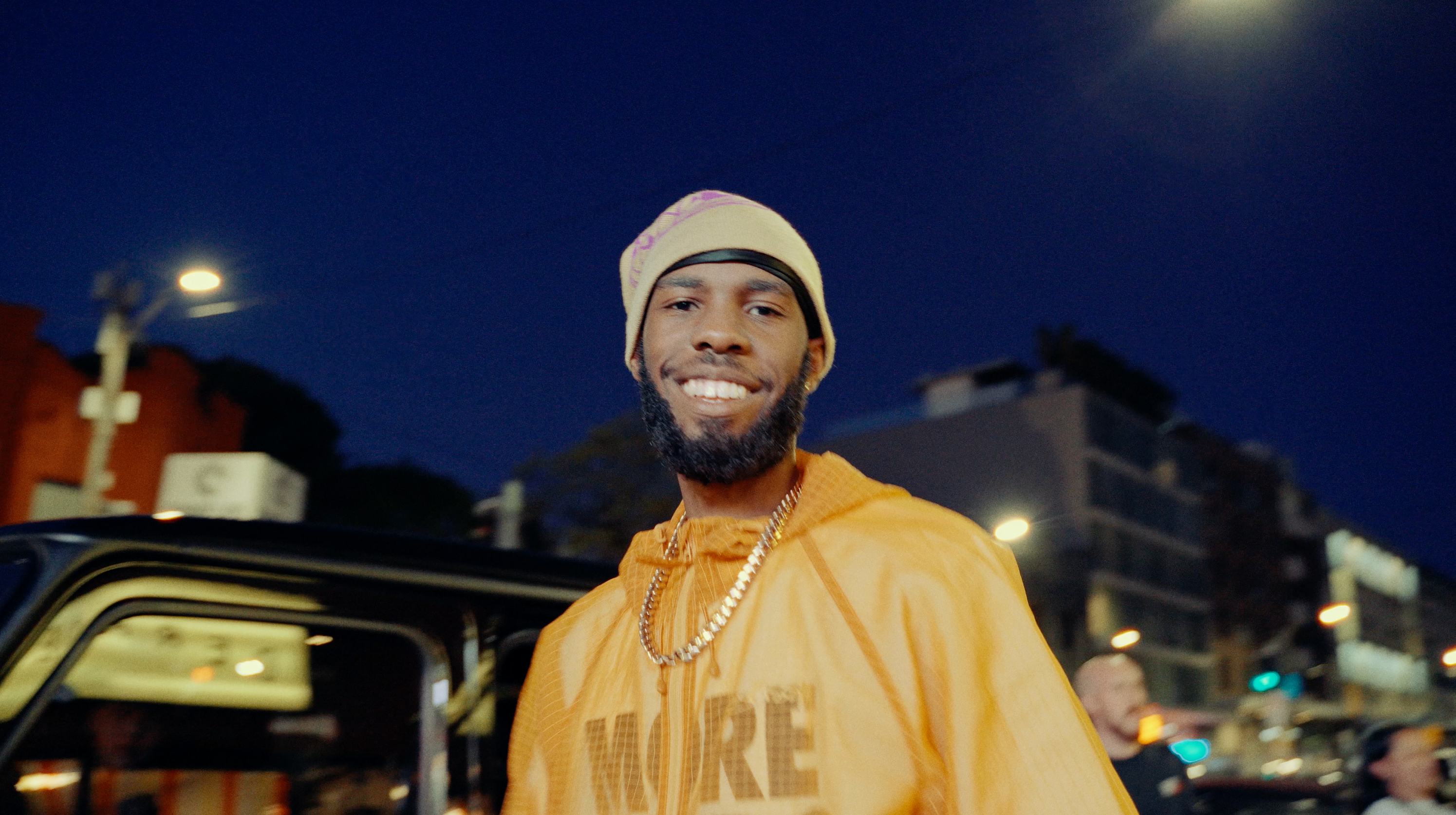 Night portrait of UK rapper Sainté in a yellow jacket and cream beanie, wearing gold chains outside a car at street level