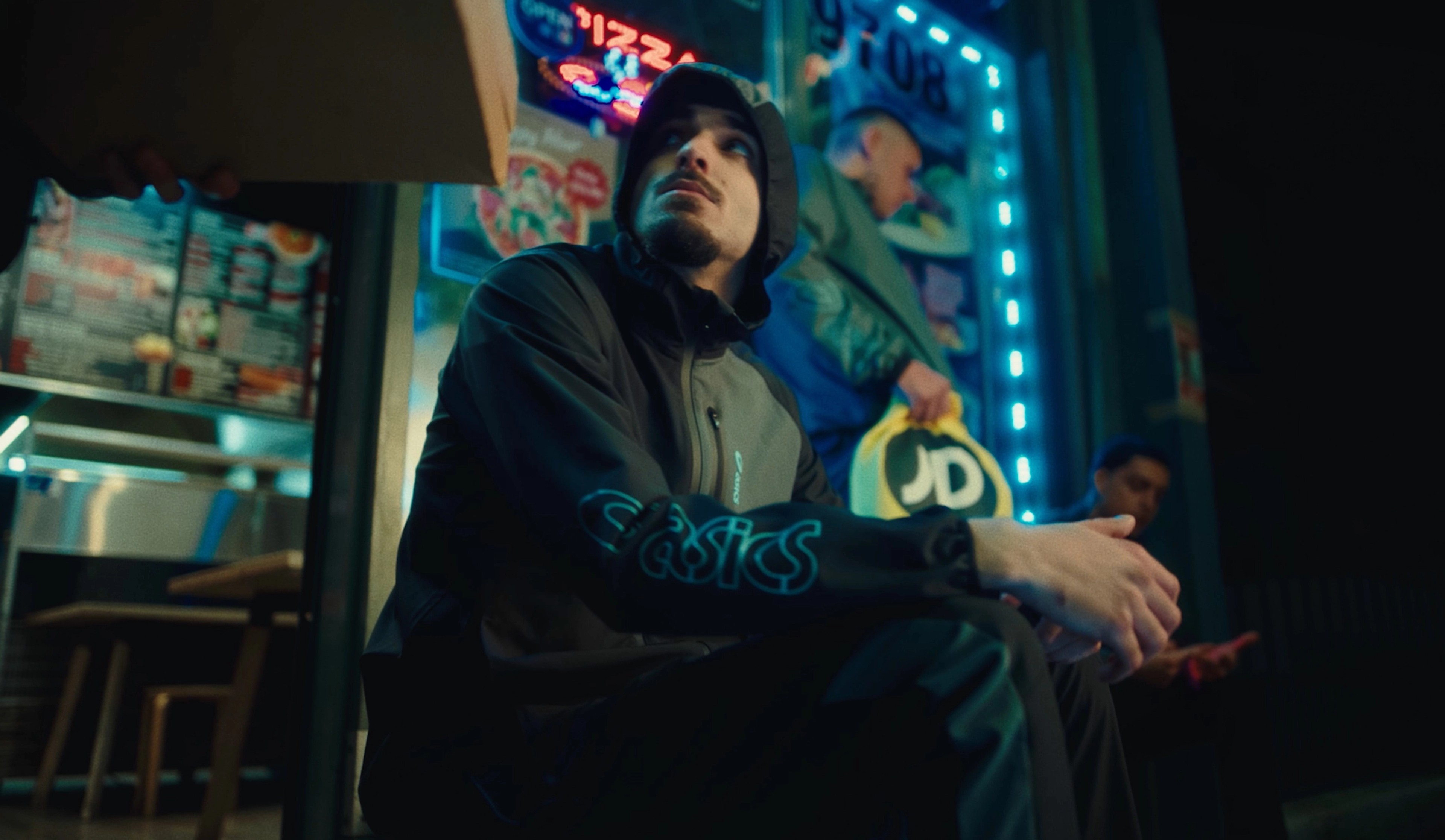 A young man in an ASICS jacket sits outside a neon-lit pizza shop at night with a JD Sports bag nearby