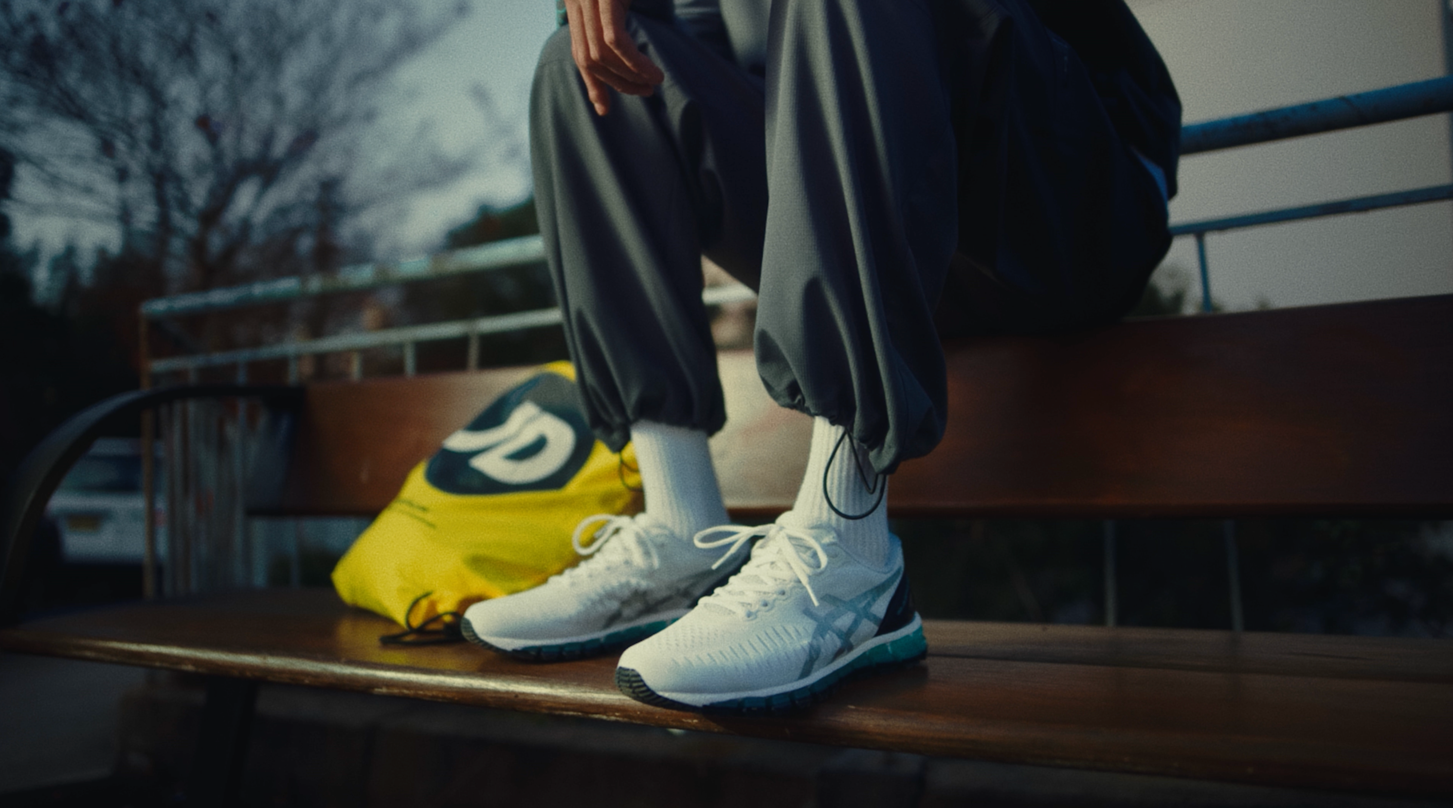 Seated figure in grey trackpants and white ASICS GEL-Quantum 360 trainers on a wooden bench, with a yellow JD Sports drawstring bag at their feet