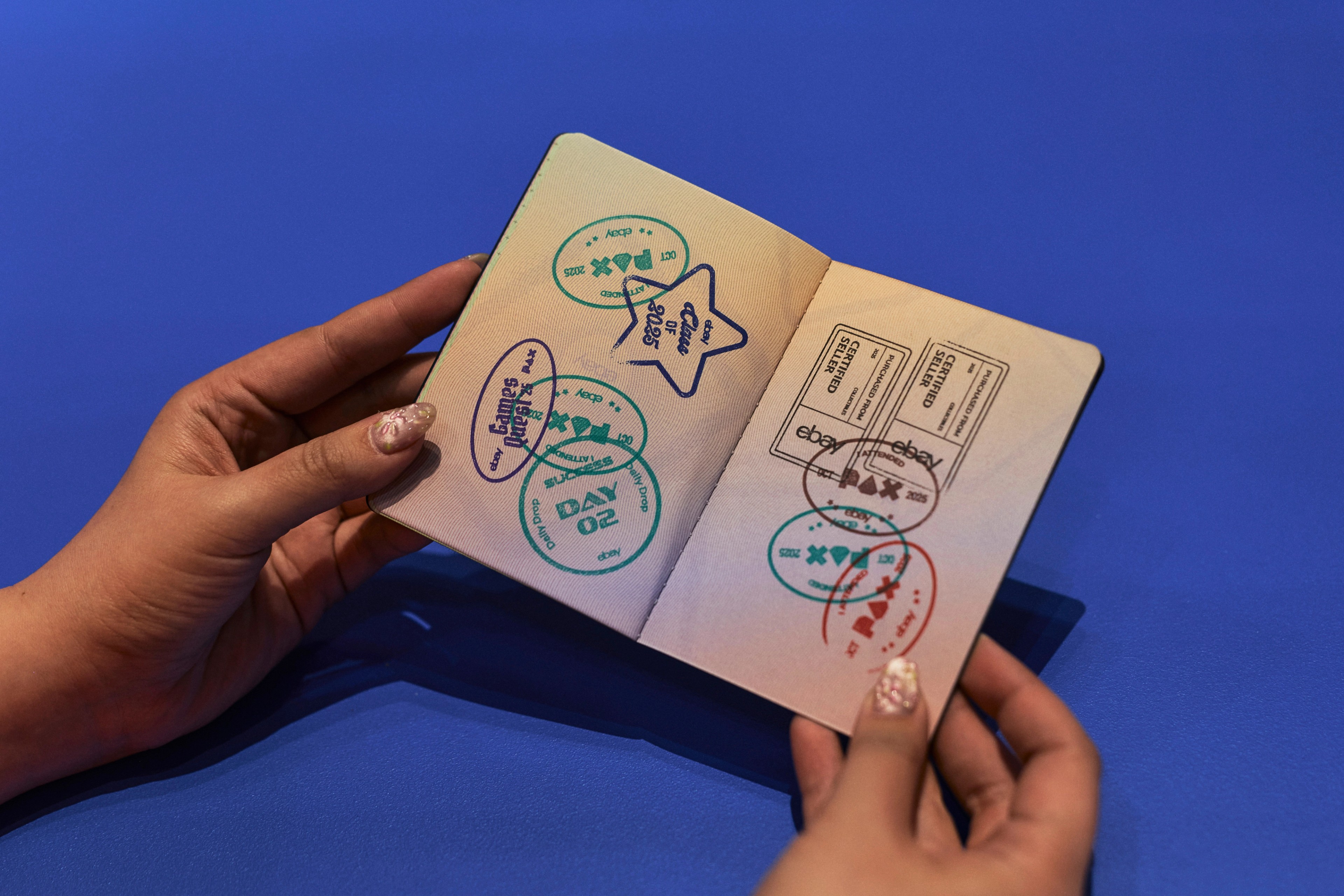 Close-up of hands holding an open eBay Passport booklet stamped with ‘Class of 2025’, ‘First Trade’ and other collector badges against a blue backdrop