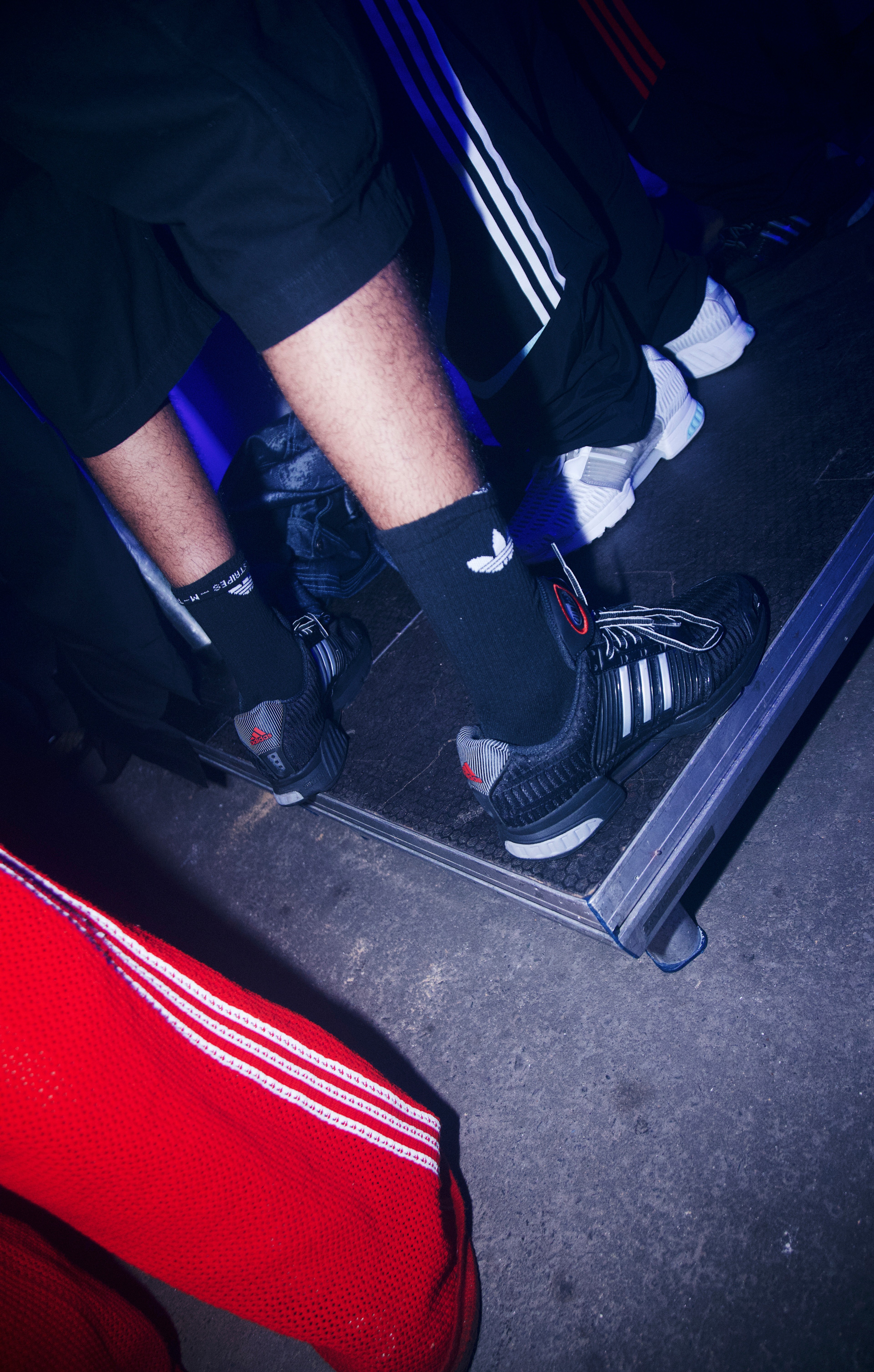 Detail shot of black adidas CLIMACOOL sneakers and three-stripe trackpants on the floor beside a red banner inside the tunnel venue