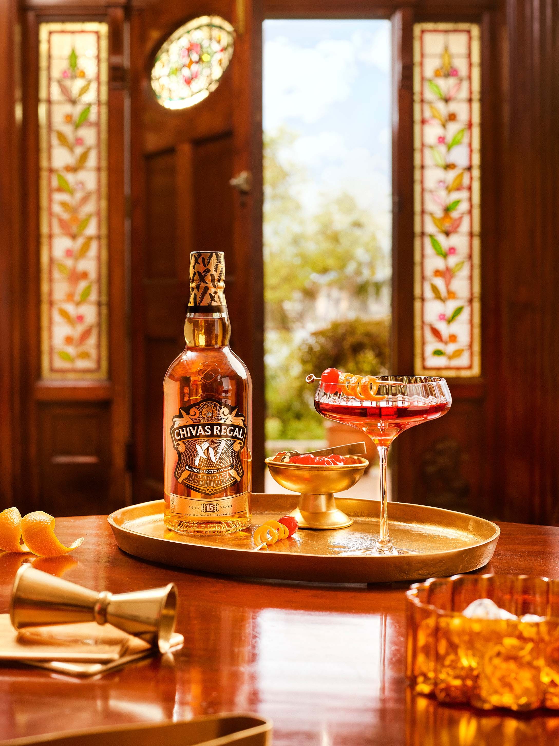 A Chivas Regal XV bottle and a red cocktail garnished with orange peel sit on a gold tray, with a jigger and cherries on a wooden table, against stained glass doors.