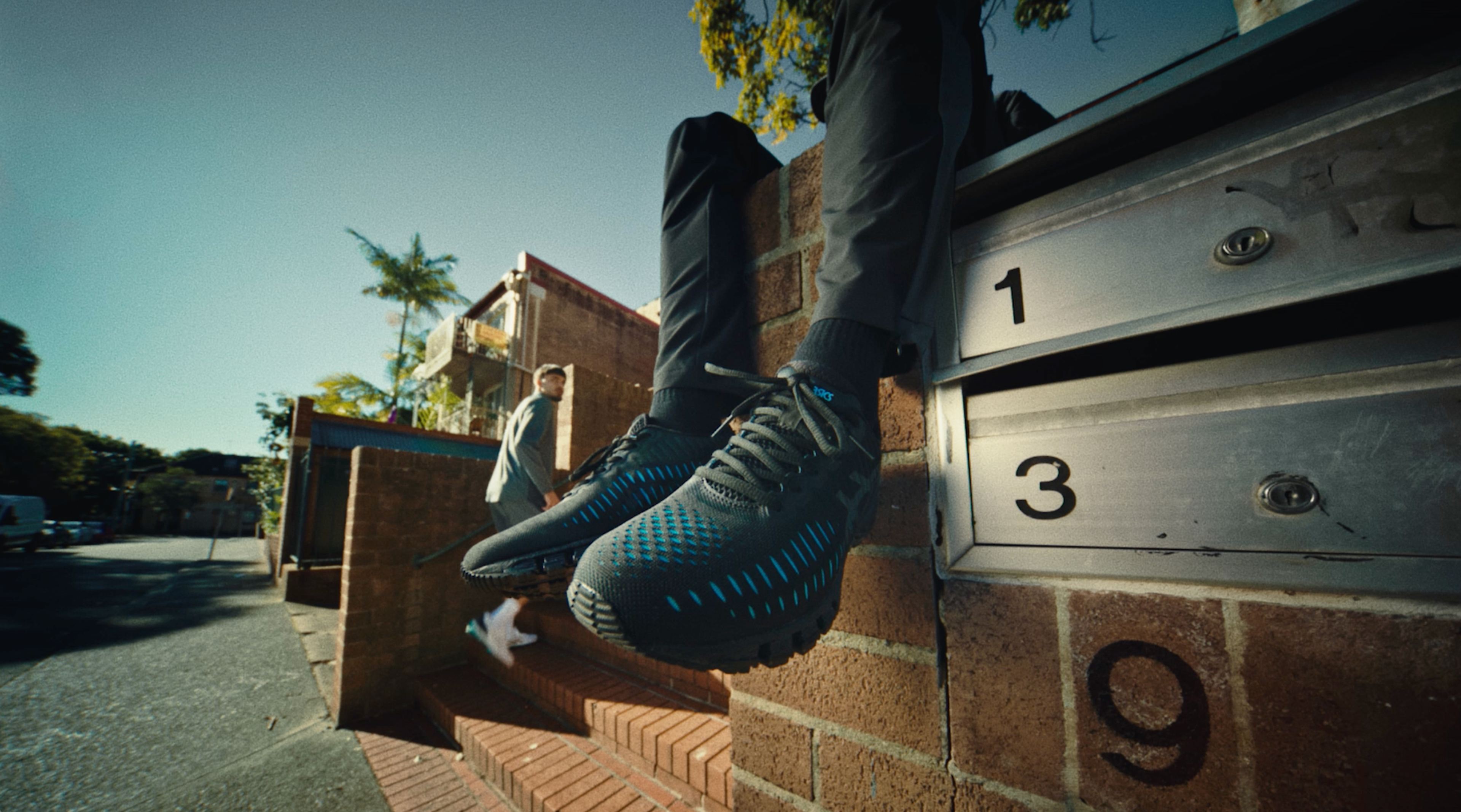 Person in black ASICS GEL-Quantum 360 shoes sitting on a brick wall next to a row of mailboxes on a sunny suburban street