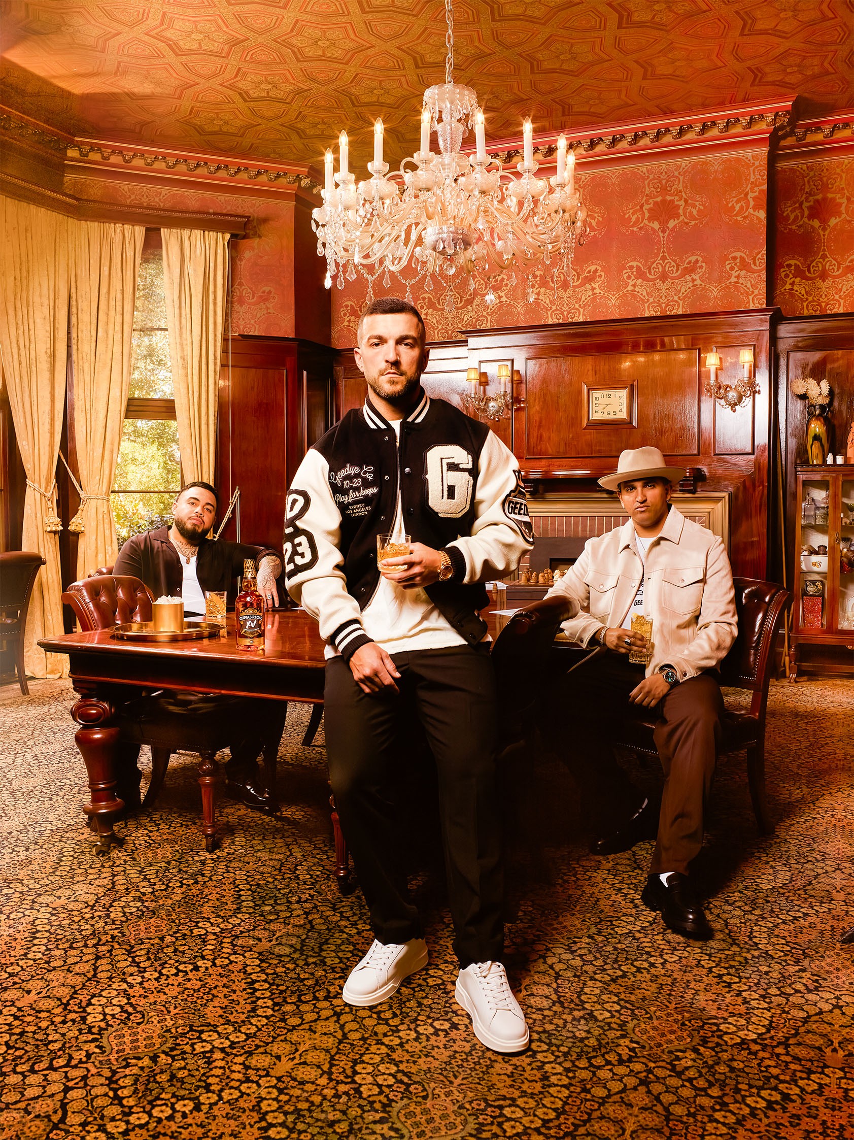 Three men, one standing in a letterman jacket and two seated, holding drinks in a luxurious wood-paneled room.