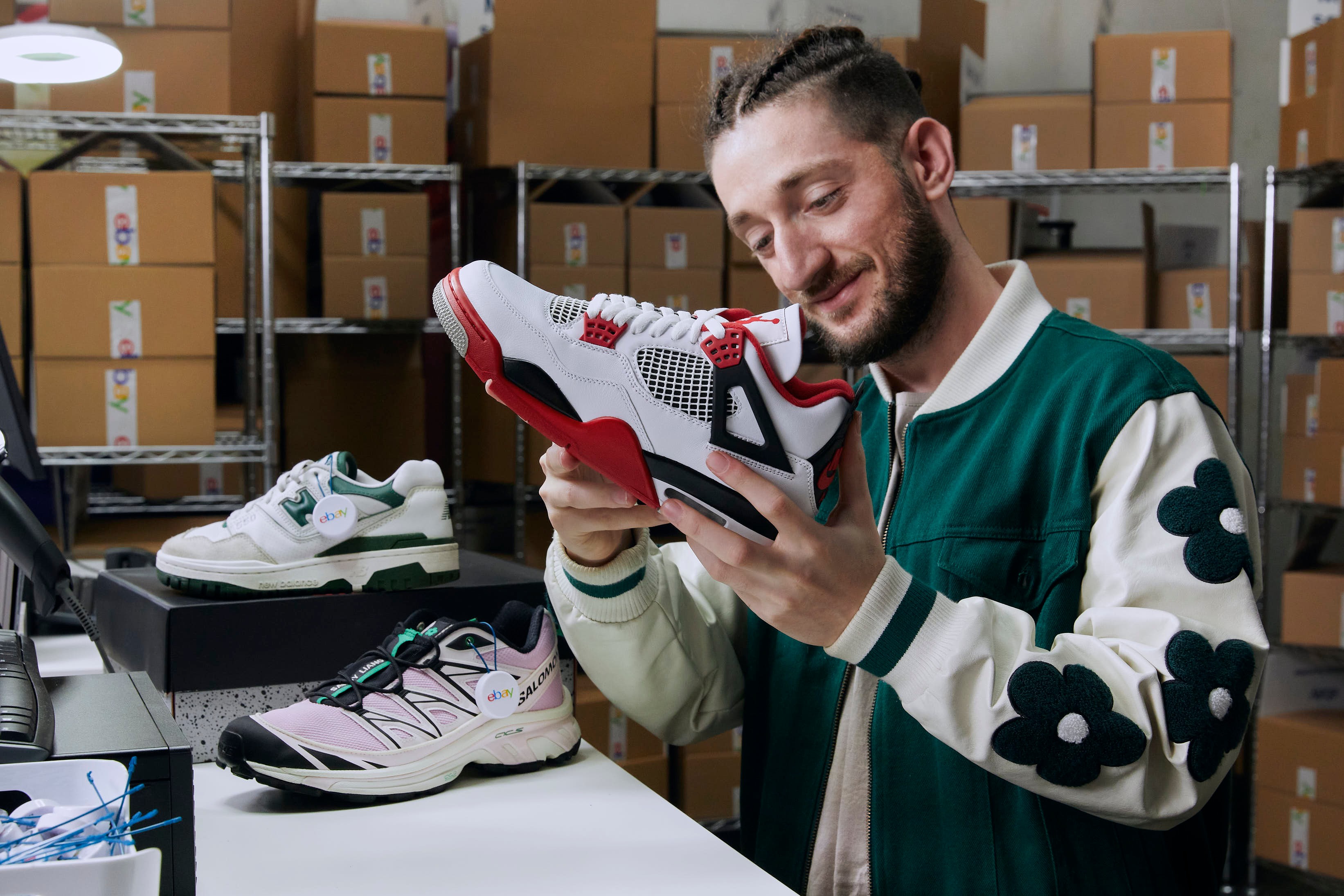 A man smiles while inspecting a red, white, and black sneaker, with other shoes and eBay-branded boxes in the background.
