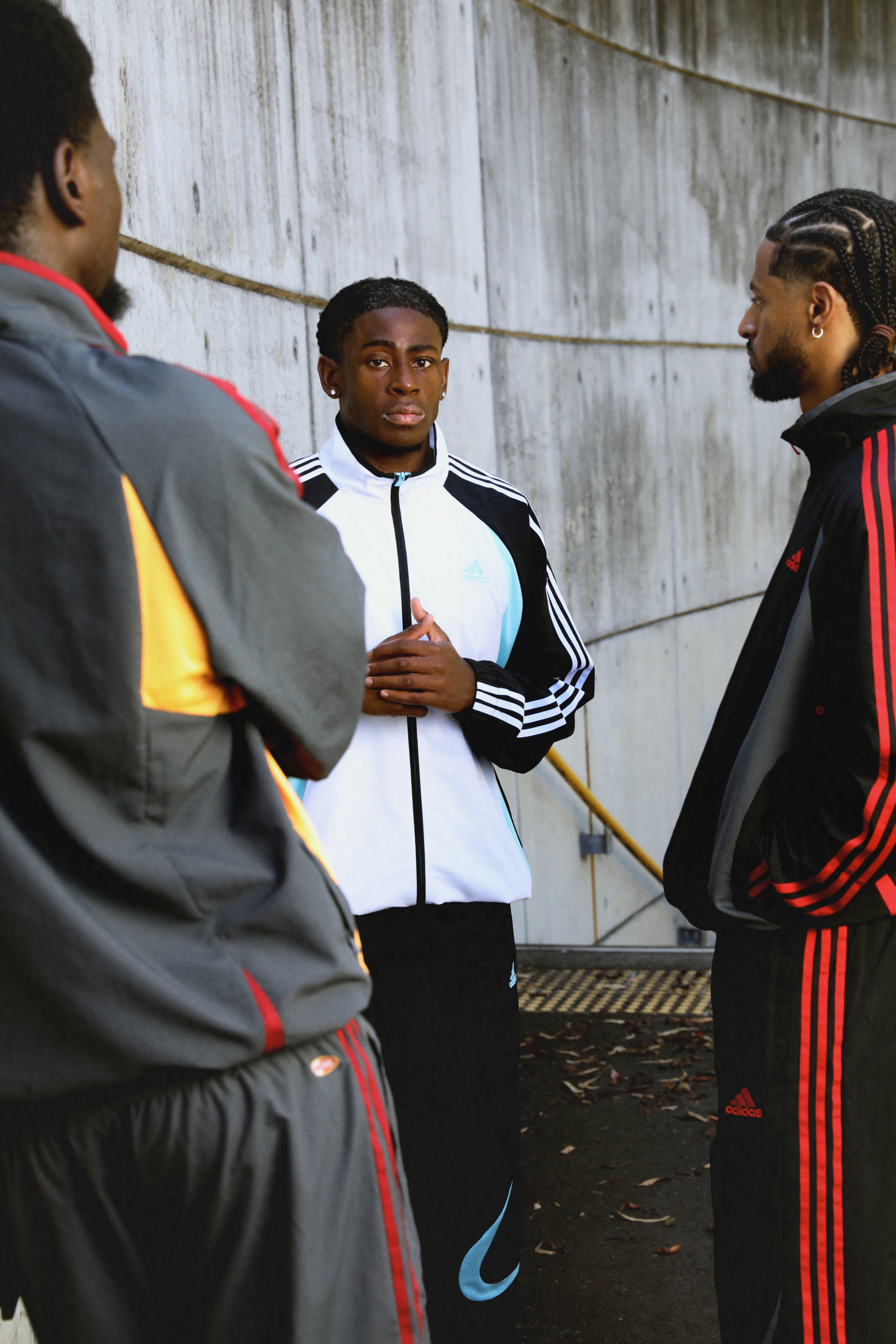 Three young men in adidas Originals tracksuits — red, white with blue accents, and black with red stripes — standing together against a concrete wall