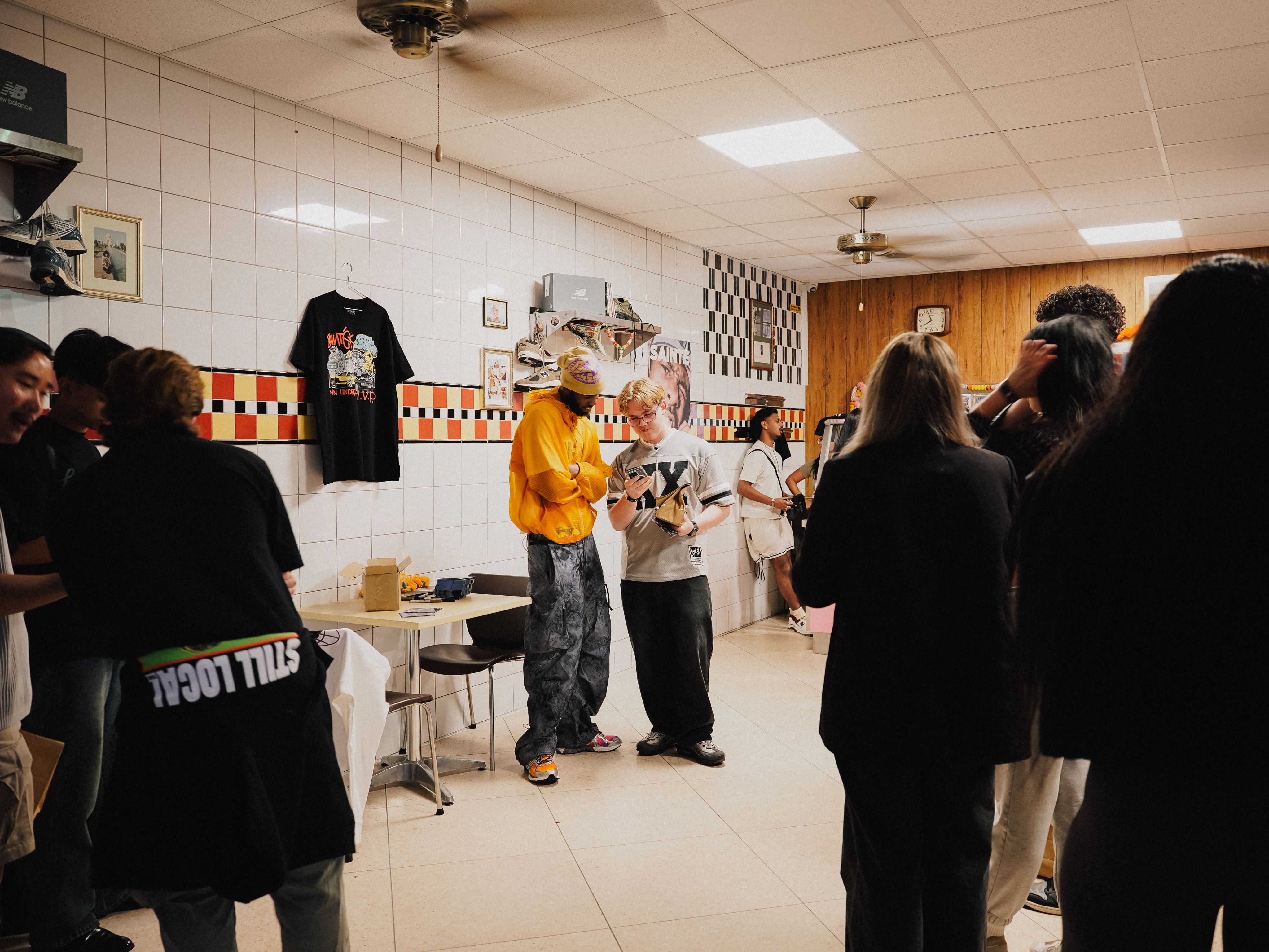 Crowd gathered inside Derrels restaurant with a black-and-white checkered floor, fans in patterned jerseys watching Sainté in a yellow jacket