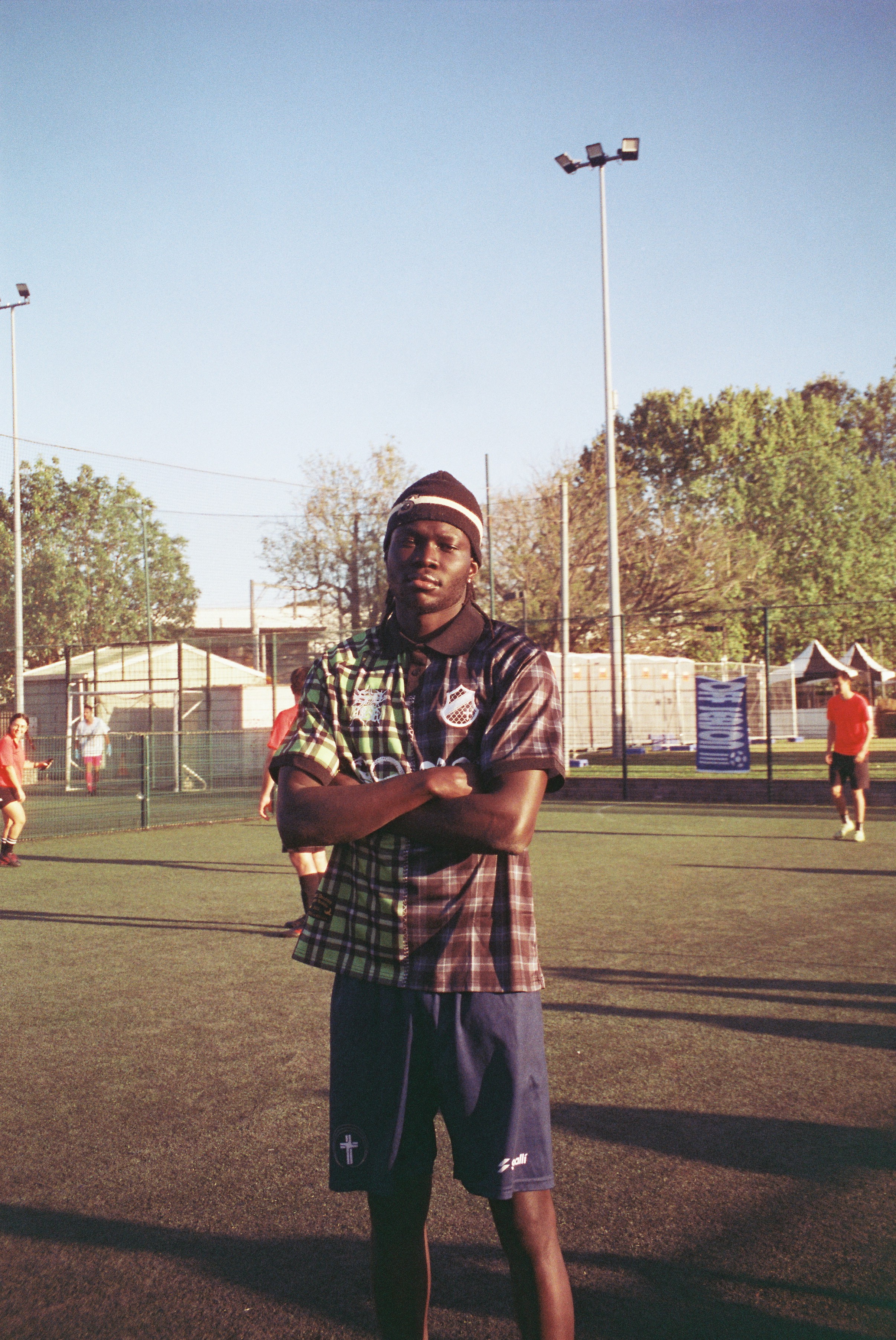 Player portrait in a red and black plaid shirt standing on a football pitch with arms crossed