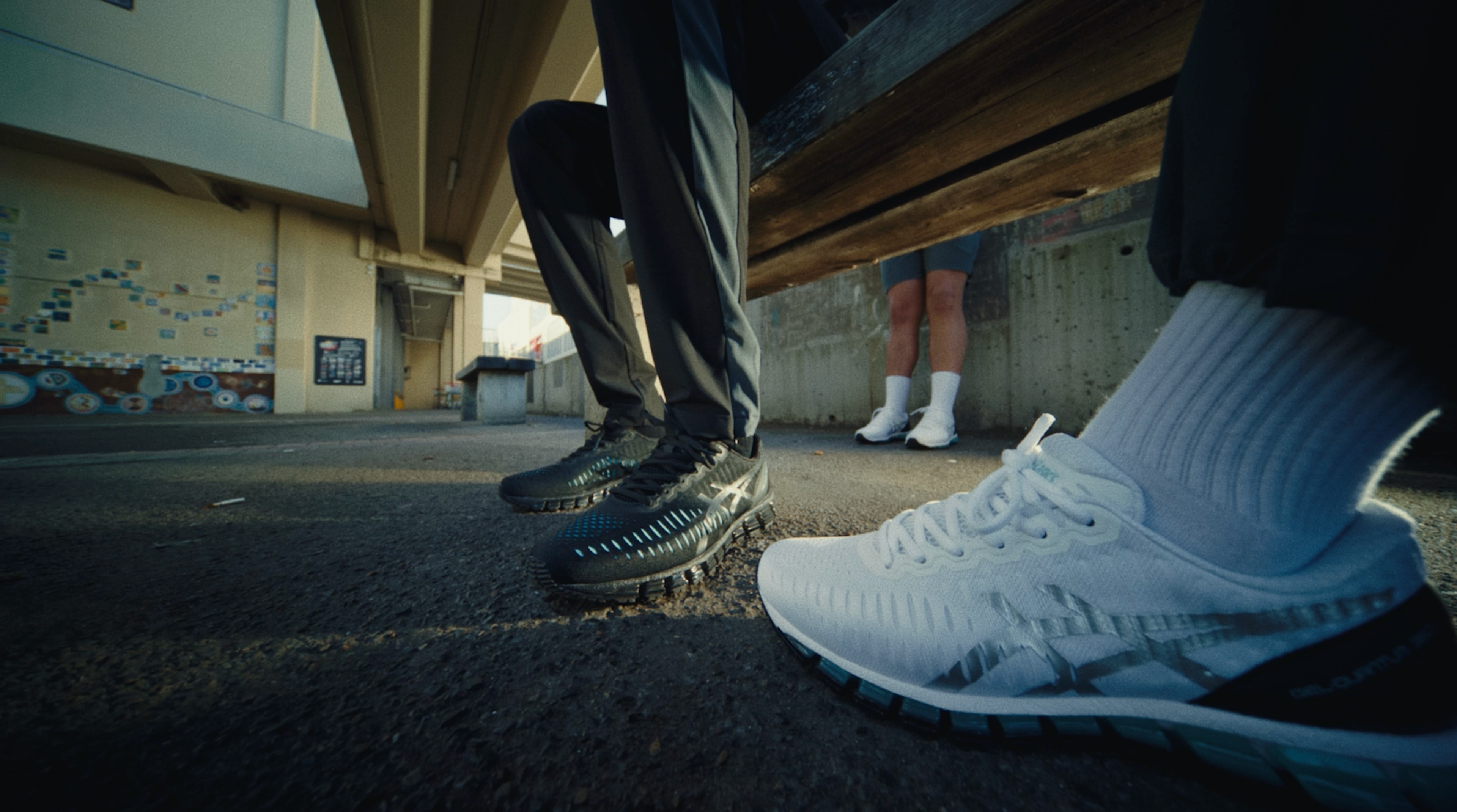 Low-angle shot of two pairs of ASICS GEL-Quantum 360 shoes — black and white — worn by figures meeting on the footpath beneath a concrete overpass