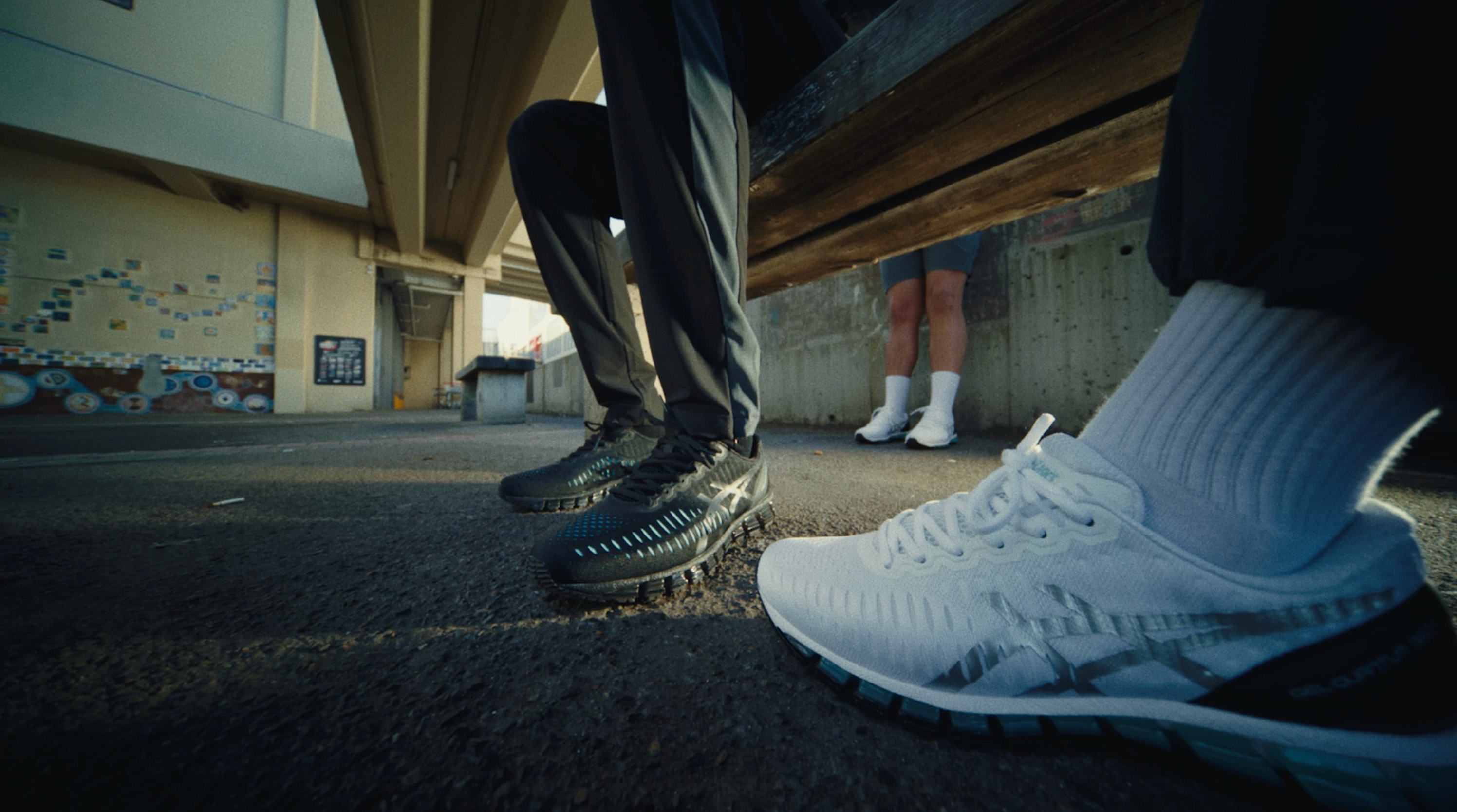 Low-angle shot of two pairs of ASICS GEL-Quantum 360 shoes — black and white — worn by figures meeting on the footpath beneath a concrete overpass