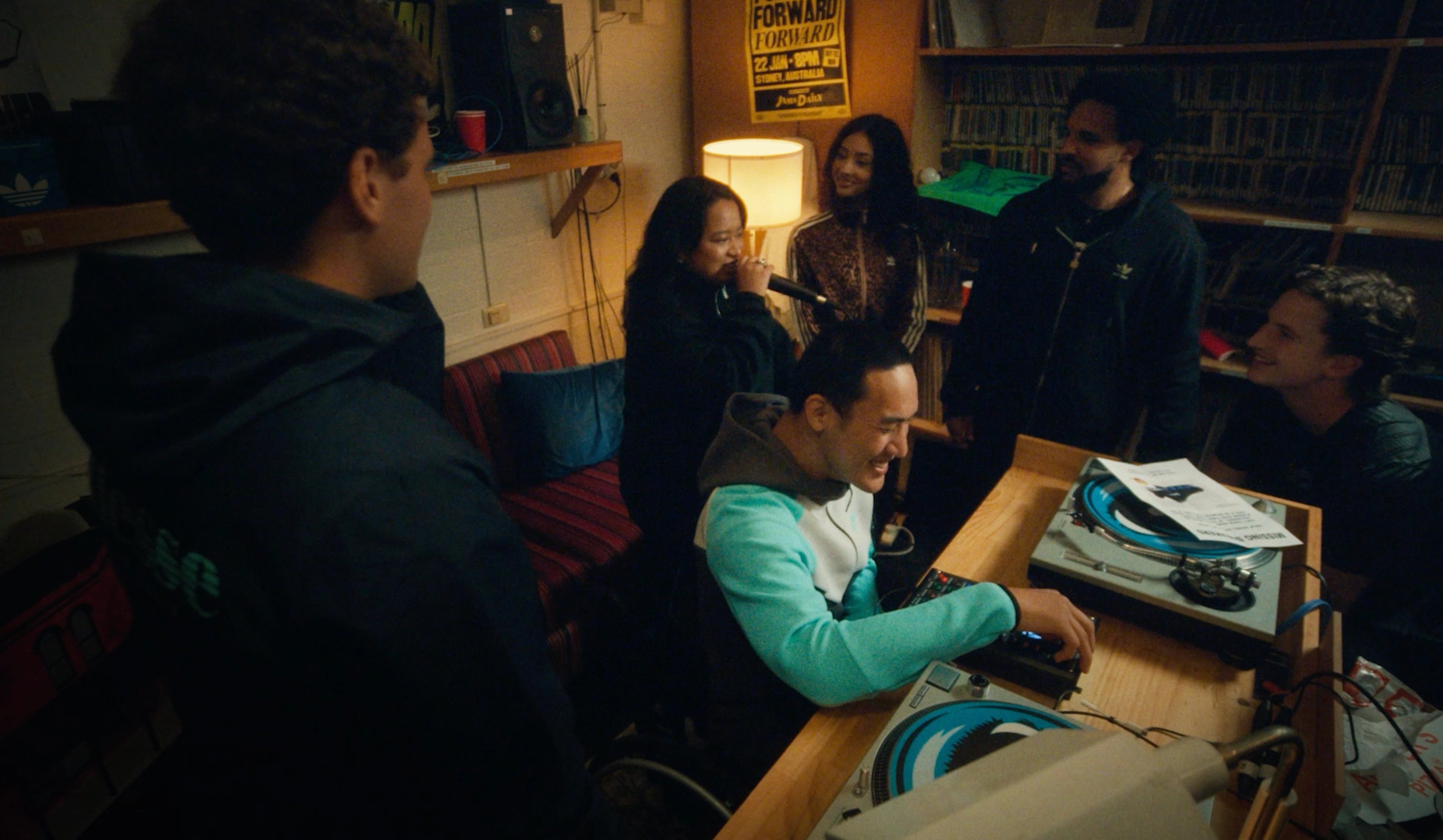 A group of friends gathered around a DJ setup in a warm, dimly lit record room with vinyl shelves and turntables
