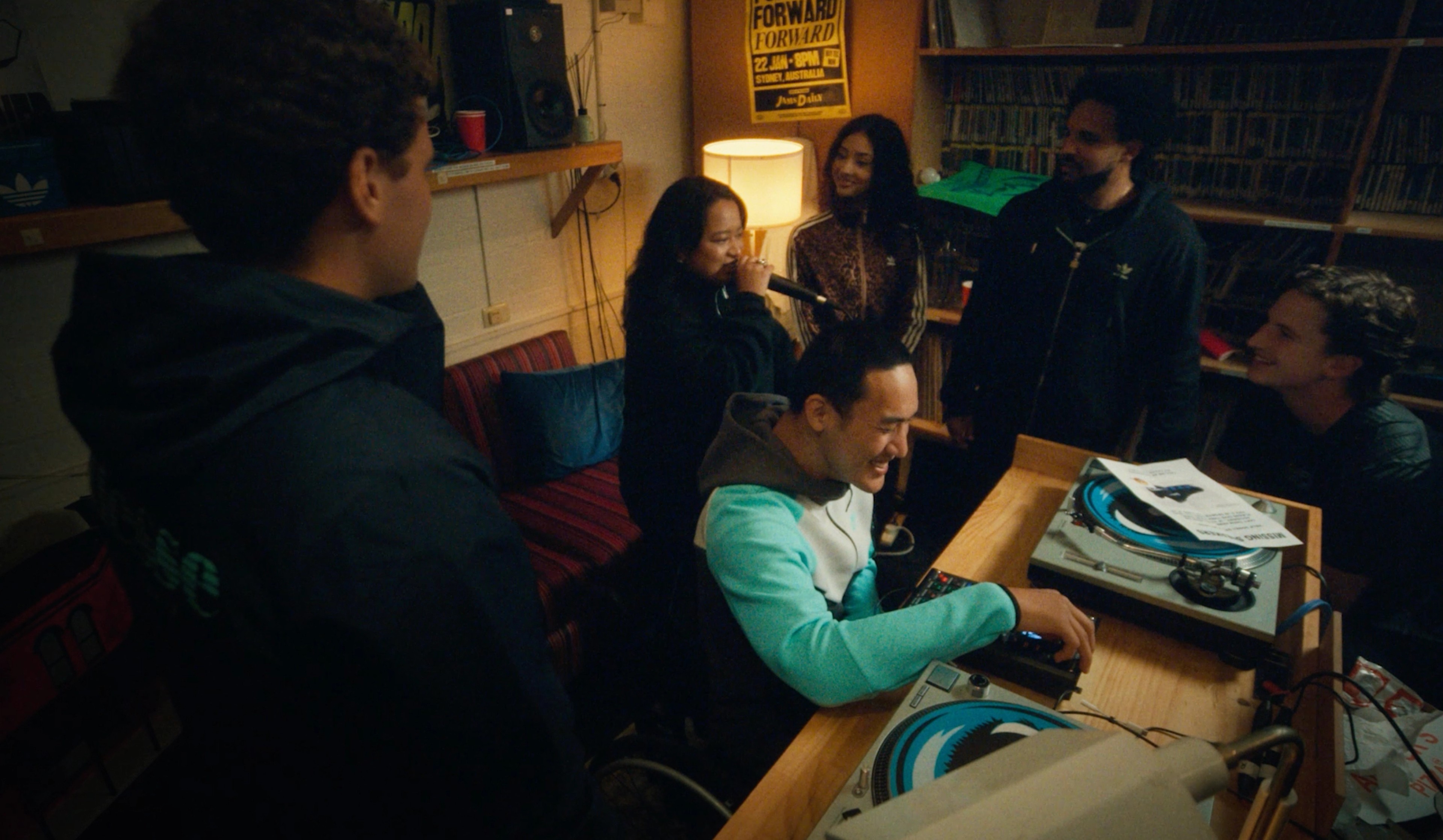 A group of friends gathered around a DJ setup in a warm, dimly lit record room with vinyl shelves and turntables