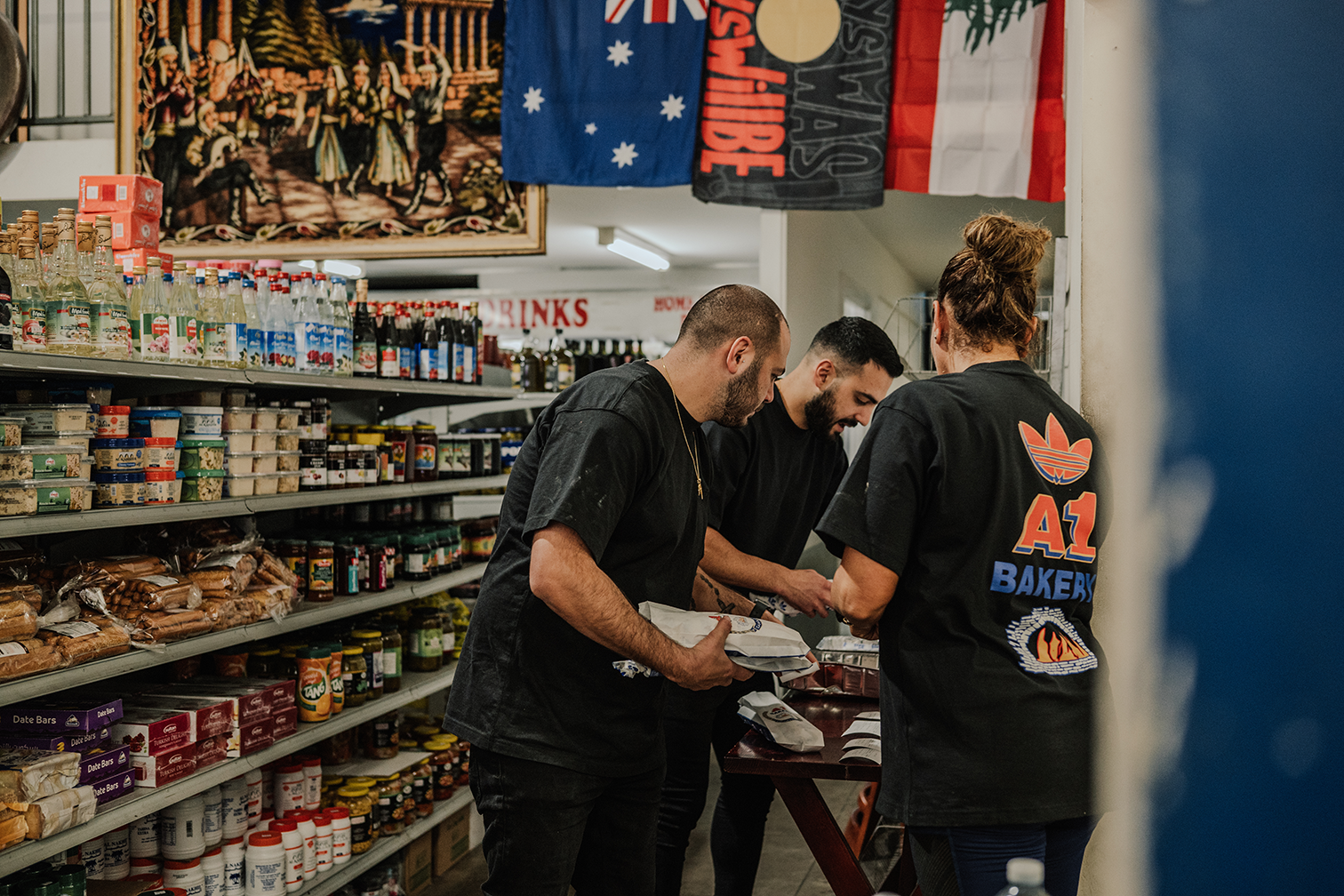Three people, including one wearing an "A1 Bakery" shirt, arrange items in a well-stocked grocery store with flags hanging overhead.