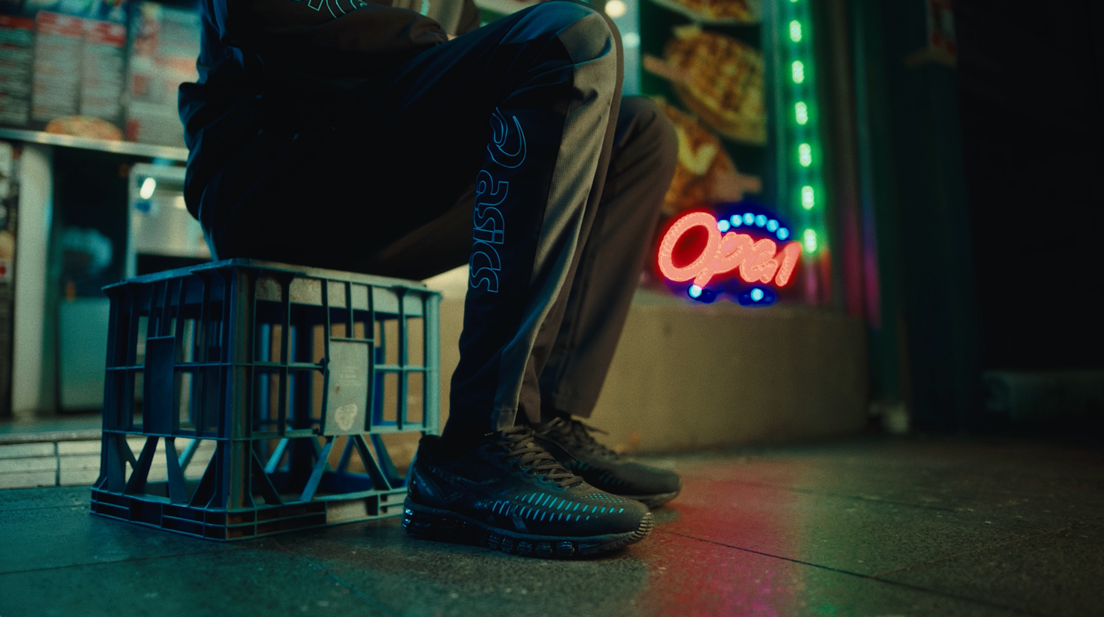 Night-time street scene with a figure perched on a milk crate in black ASICS trackpants and black GEL-Quantum 360 trainers, neon Open sign glowing in the background