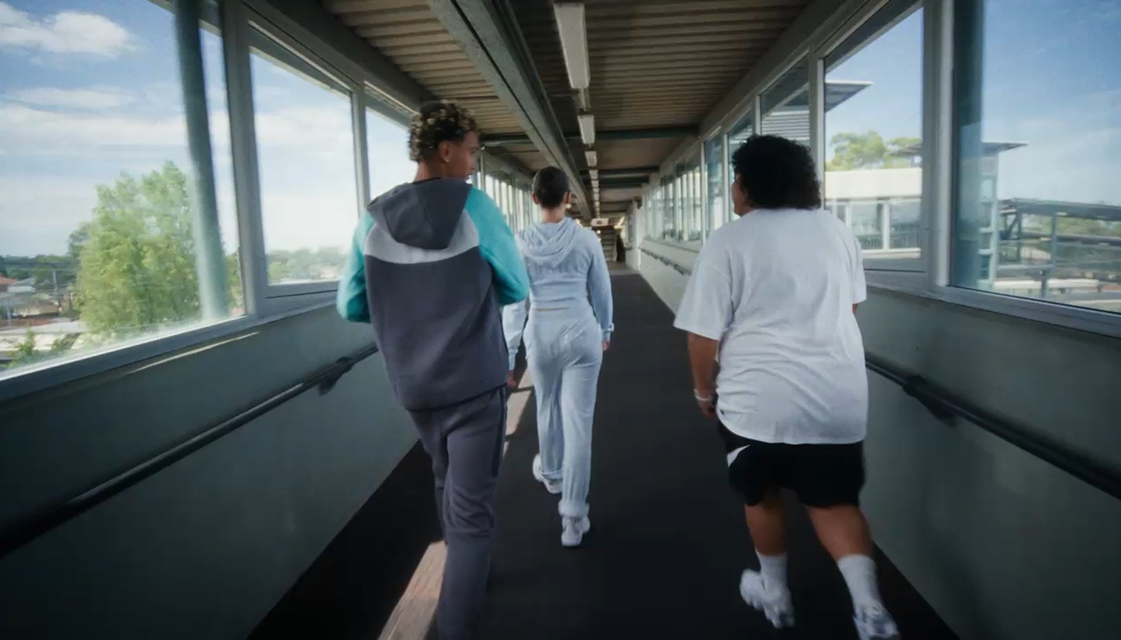 Three people walk away from the camera through a bright, windowed pedestrian bridge.