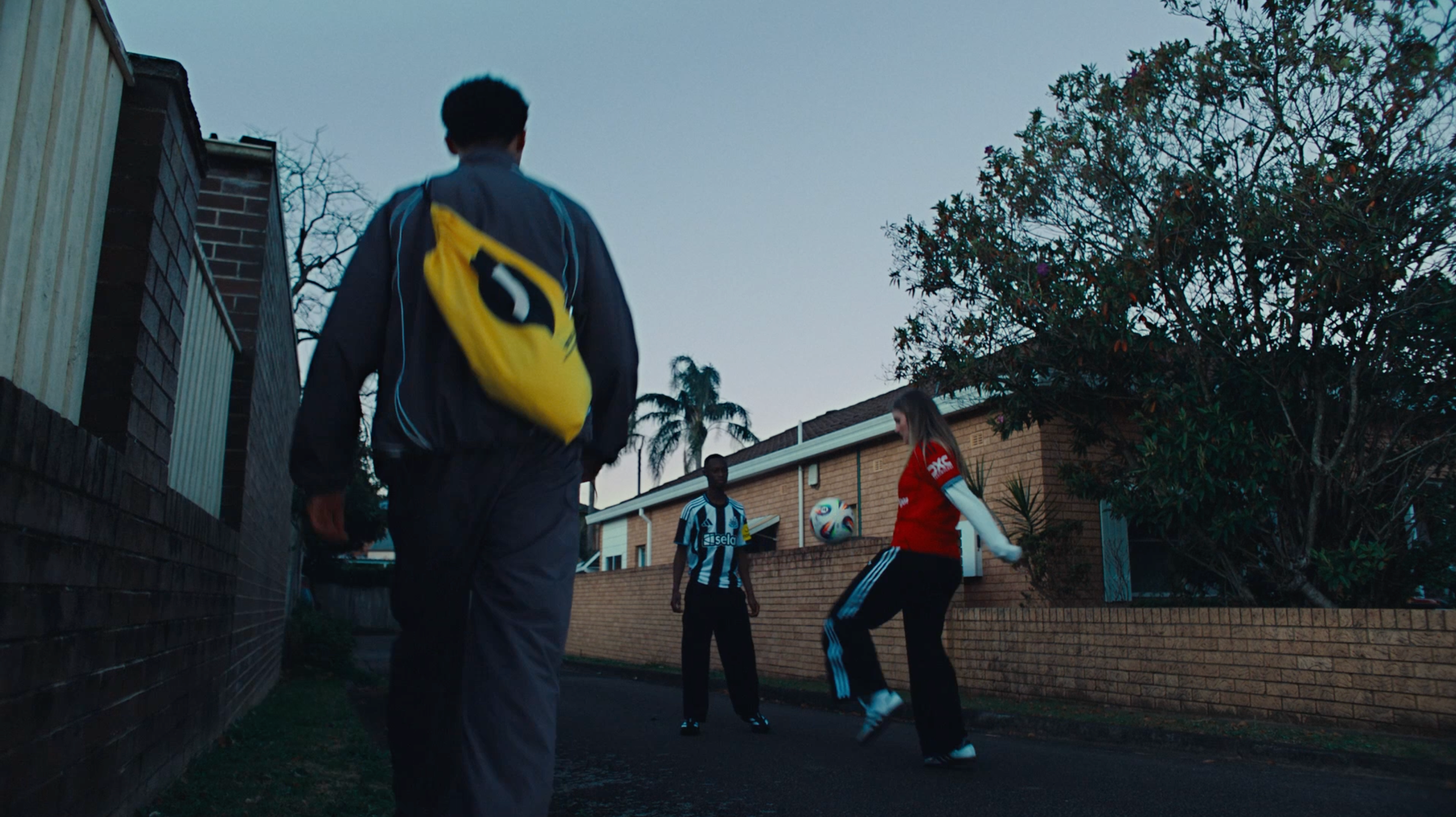 Adult in a blue jacket carrying a bright yellow backpack walking down a suburban street at dusk toward two young fans — one in a black-and-white striped jersey, the other in a red replica kit — playing with a football