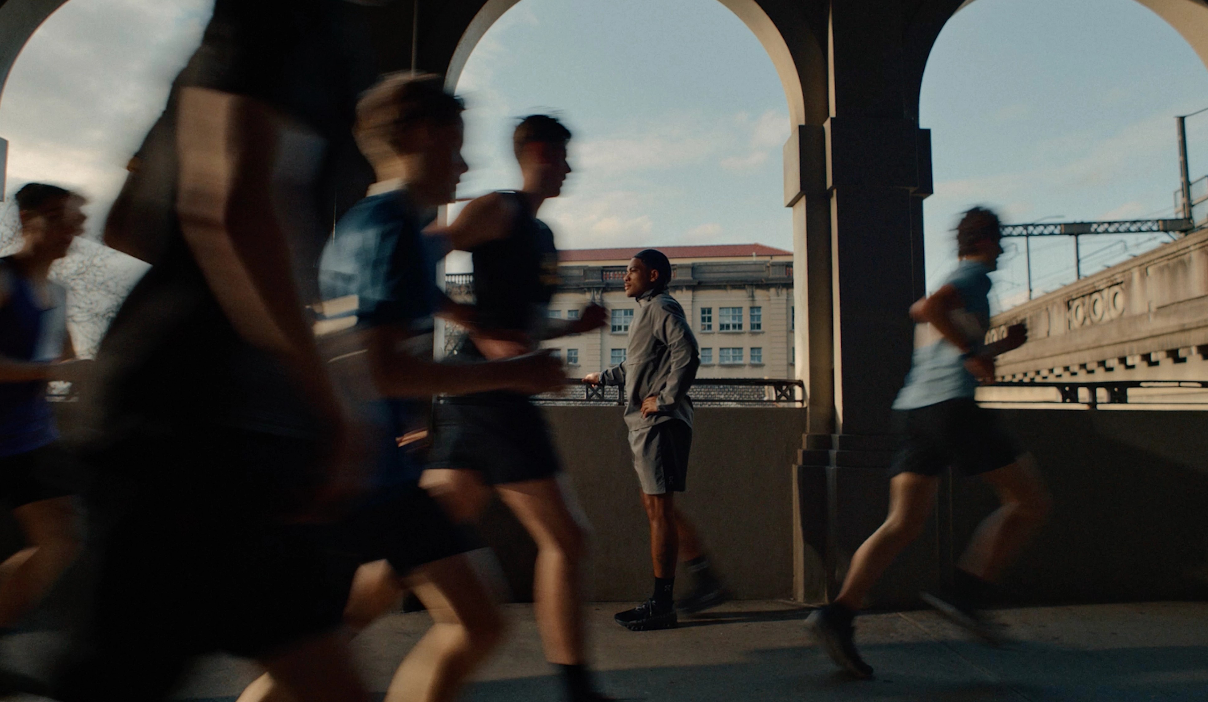 A person stands still under a bridge archway while runners blur past in motion