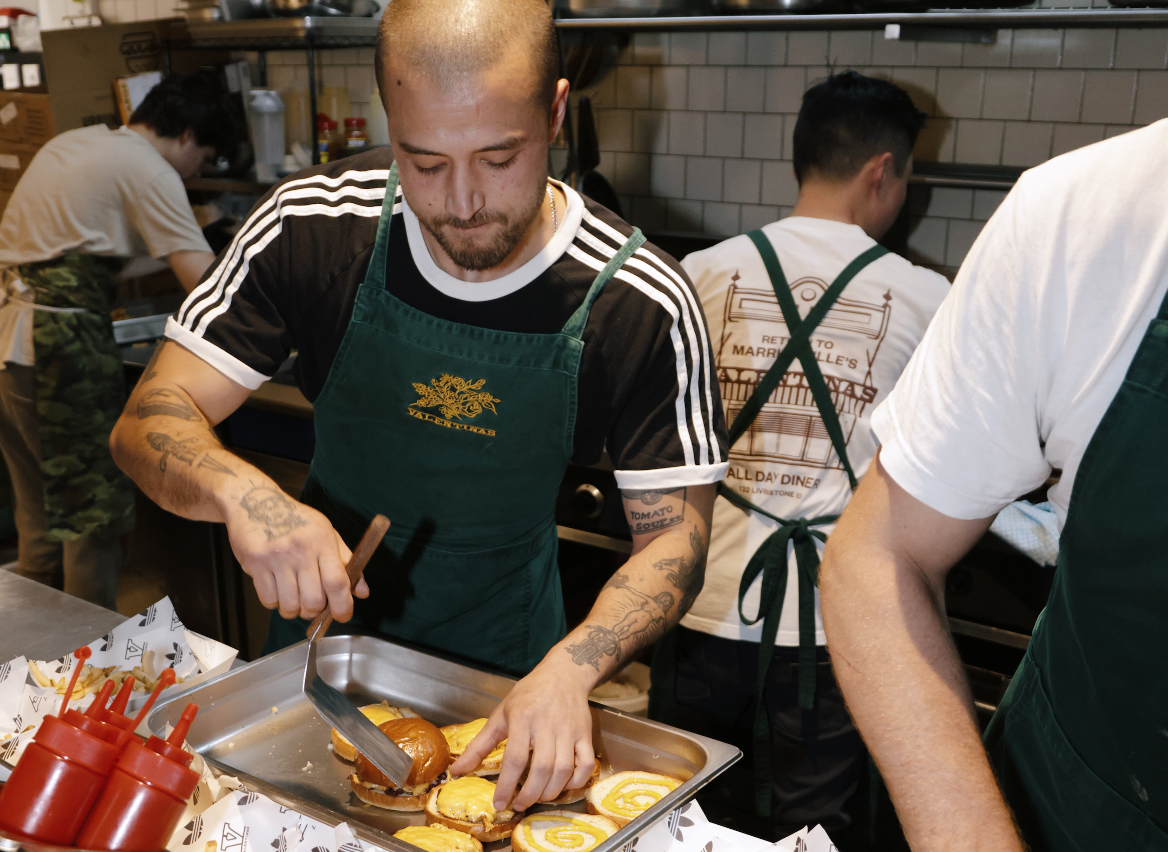 A tattooed chef in a green apron prepares burgers on a tray in a bustling kitchen.