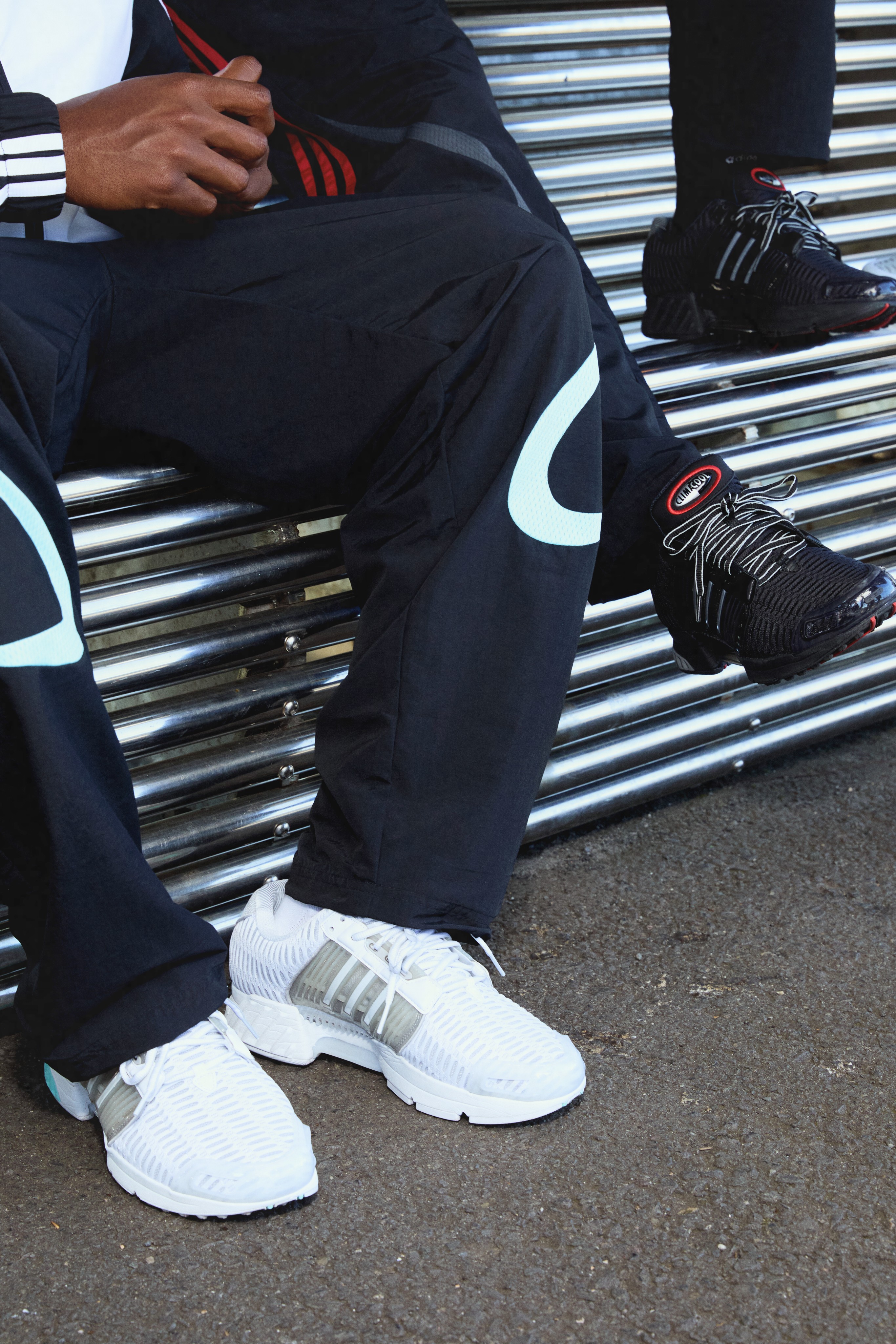 Close-up of a figure in black trackpants wearing white adidas CLIMACOOL sneakers, seated on corrugated metal steps