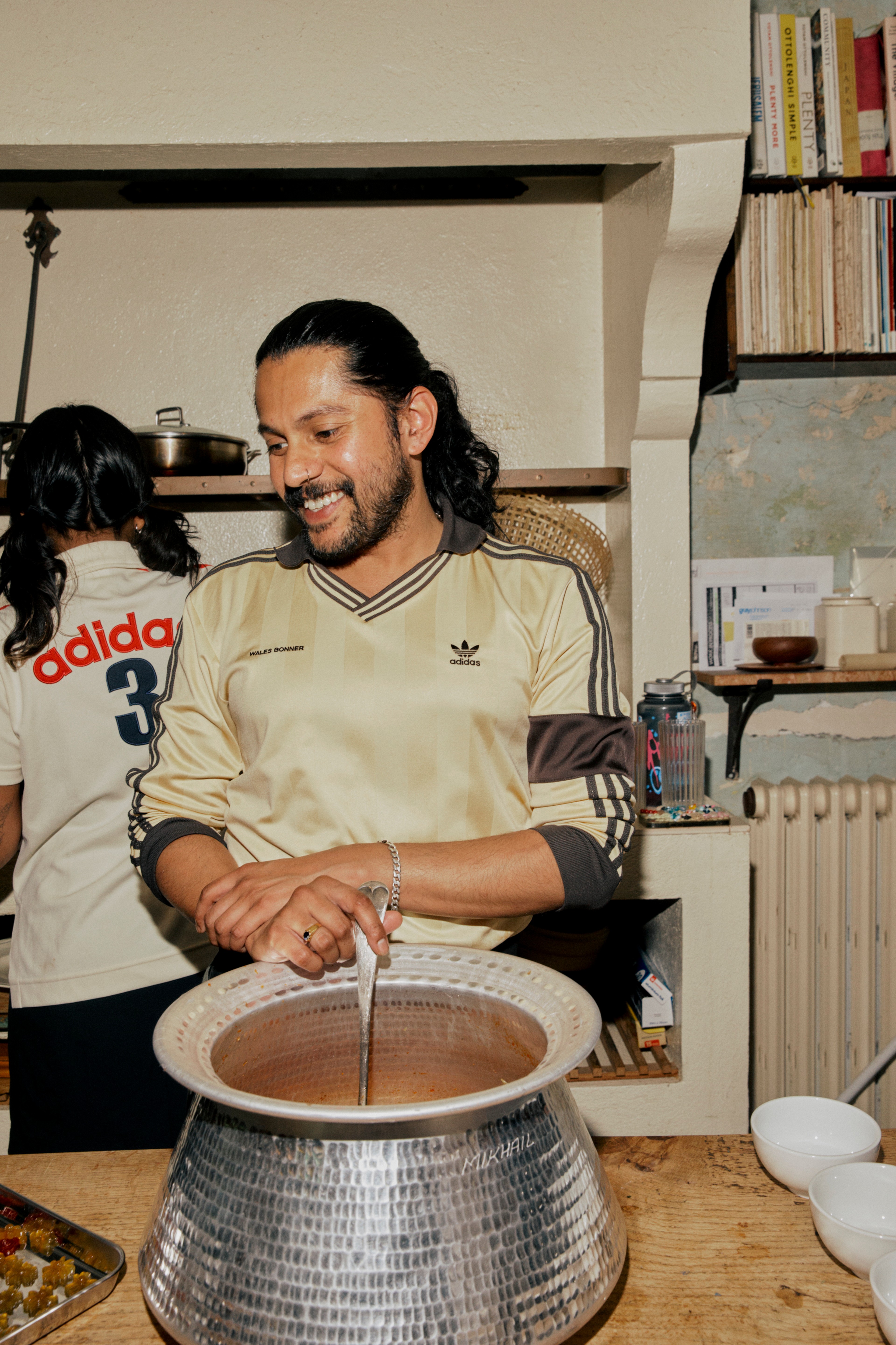 A smiling man in a beige Adidas shirt stirs a large pot of food in a kitchen.