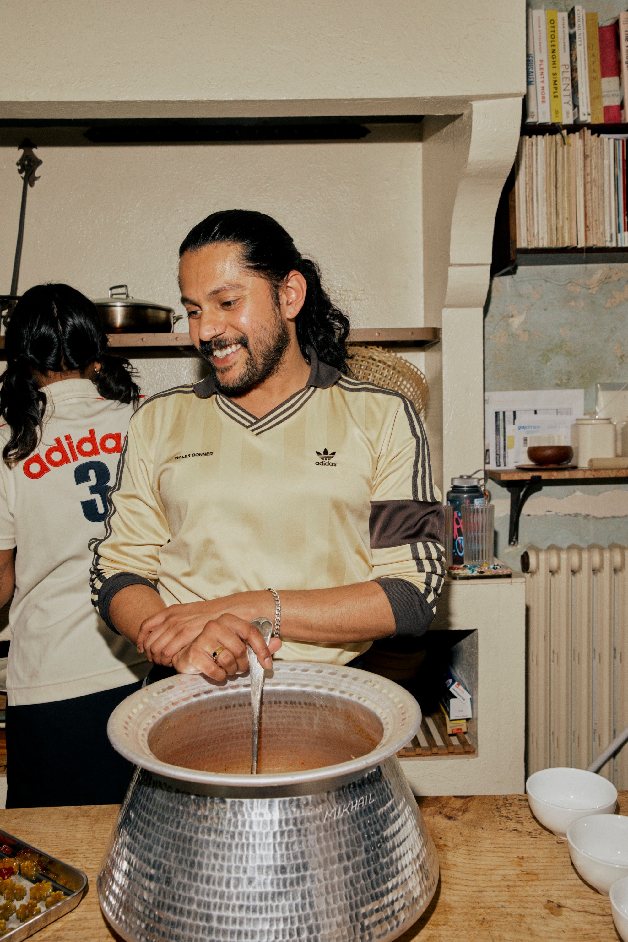 A smiling man in a beige Adidas shirt stirs a large pot of food in a kitchen.