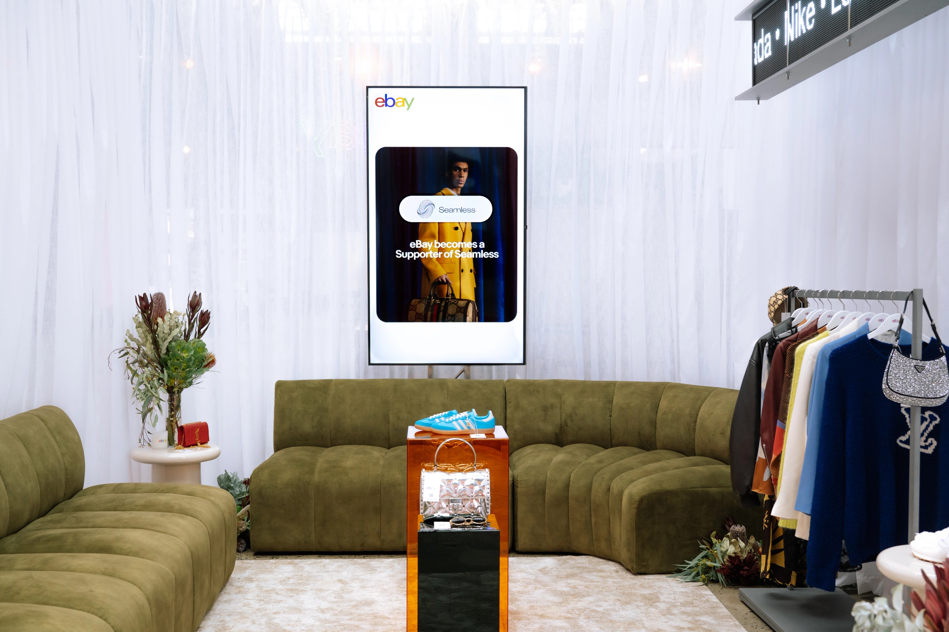 eBay-branded vertical TV display flanked by olive-green lounge seating inside the Pre-Loved Lounge