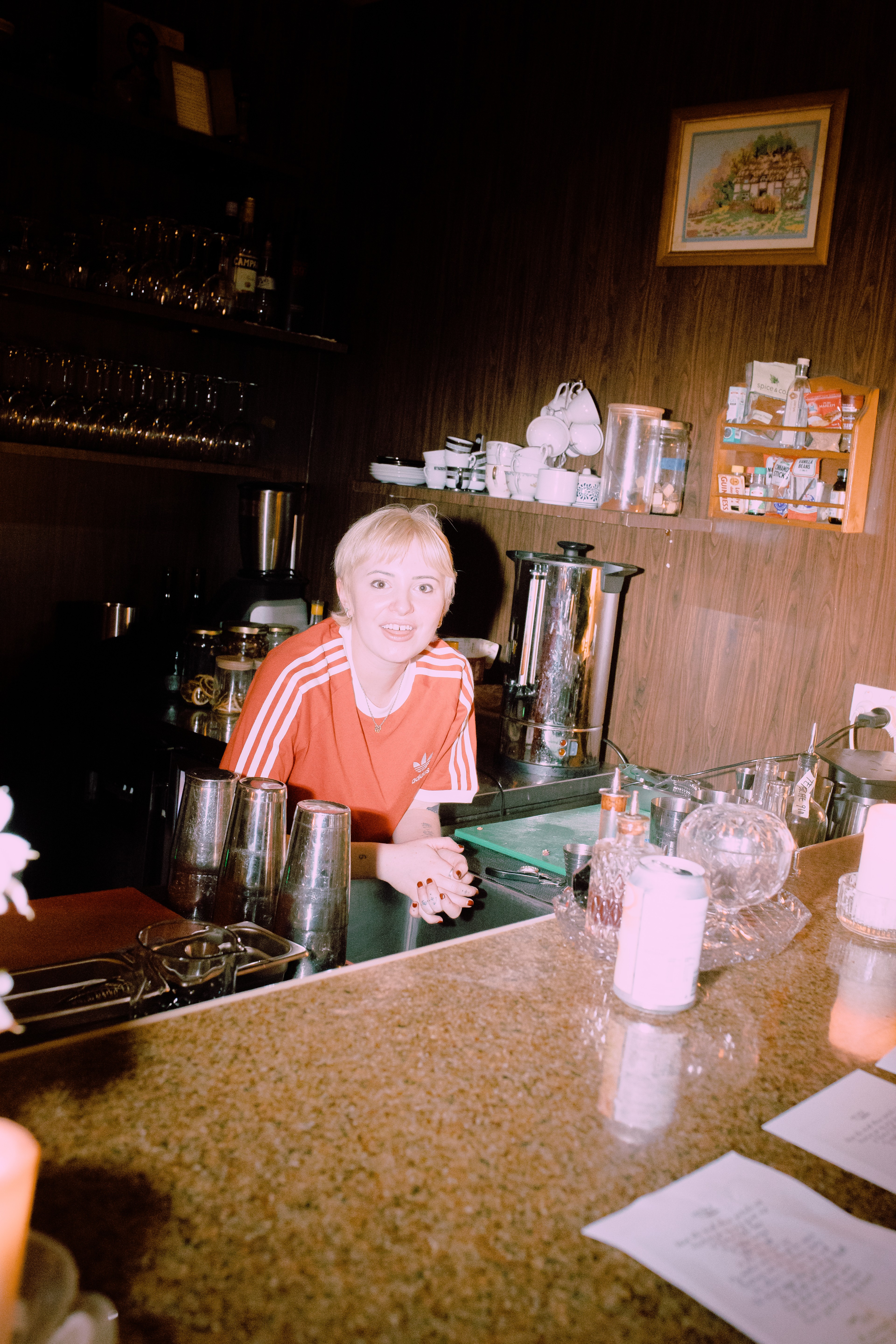A blonde woman in a red Adidas shirt smiles behind a bar counter.