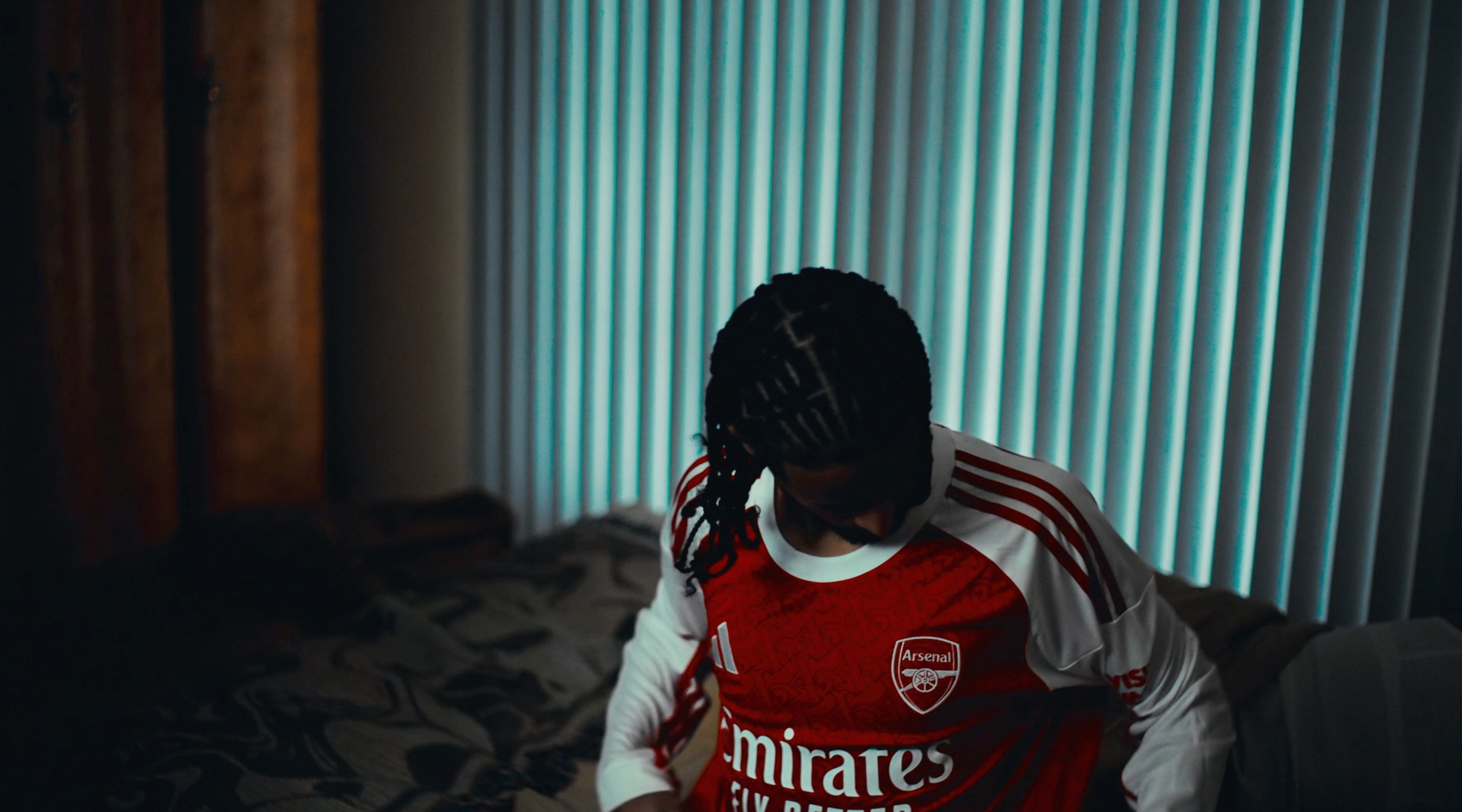 Young fan in a red Arsenal Emirates replica jersey seated inside a darkened bedroom with blue horizontal blinds behind, head tilted and looking away
