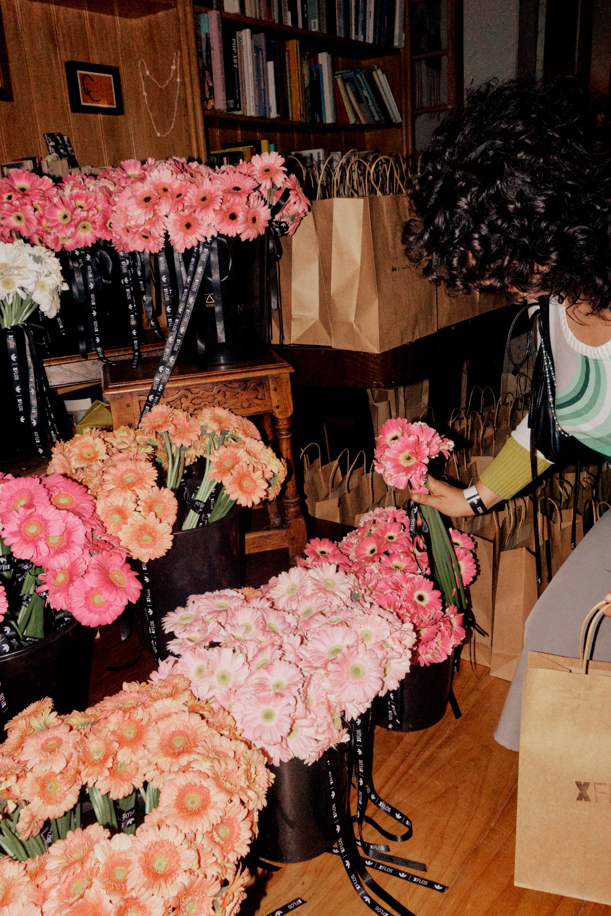A person picks a pink gerbera daisy from an array of pink and orange gerbera bouquets in a room with bookshelves and brown paper bags.