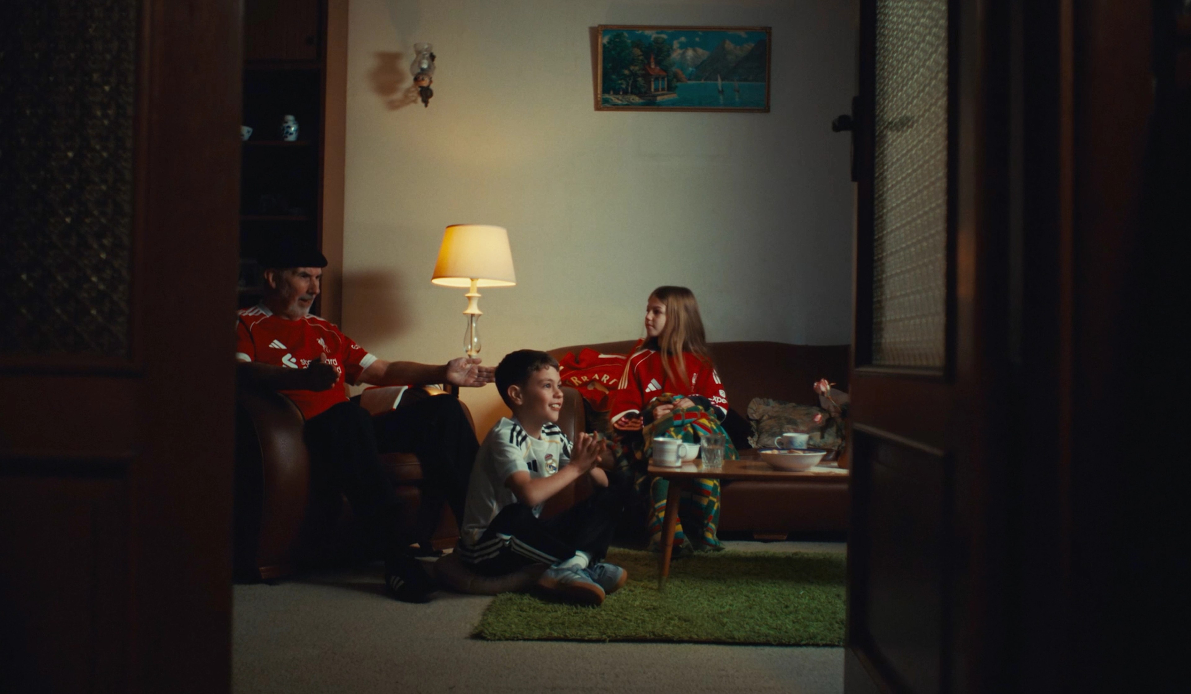 A family in football jerseys watches a match together in a warm, lamp-lit living room