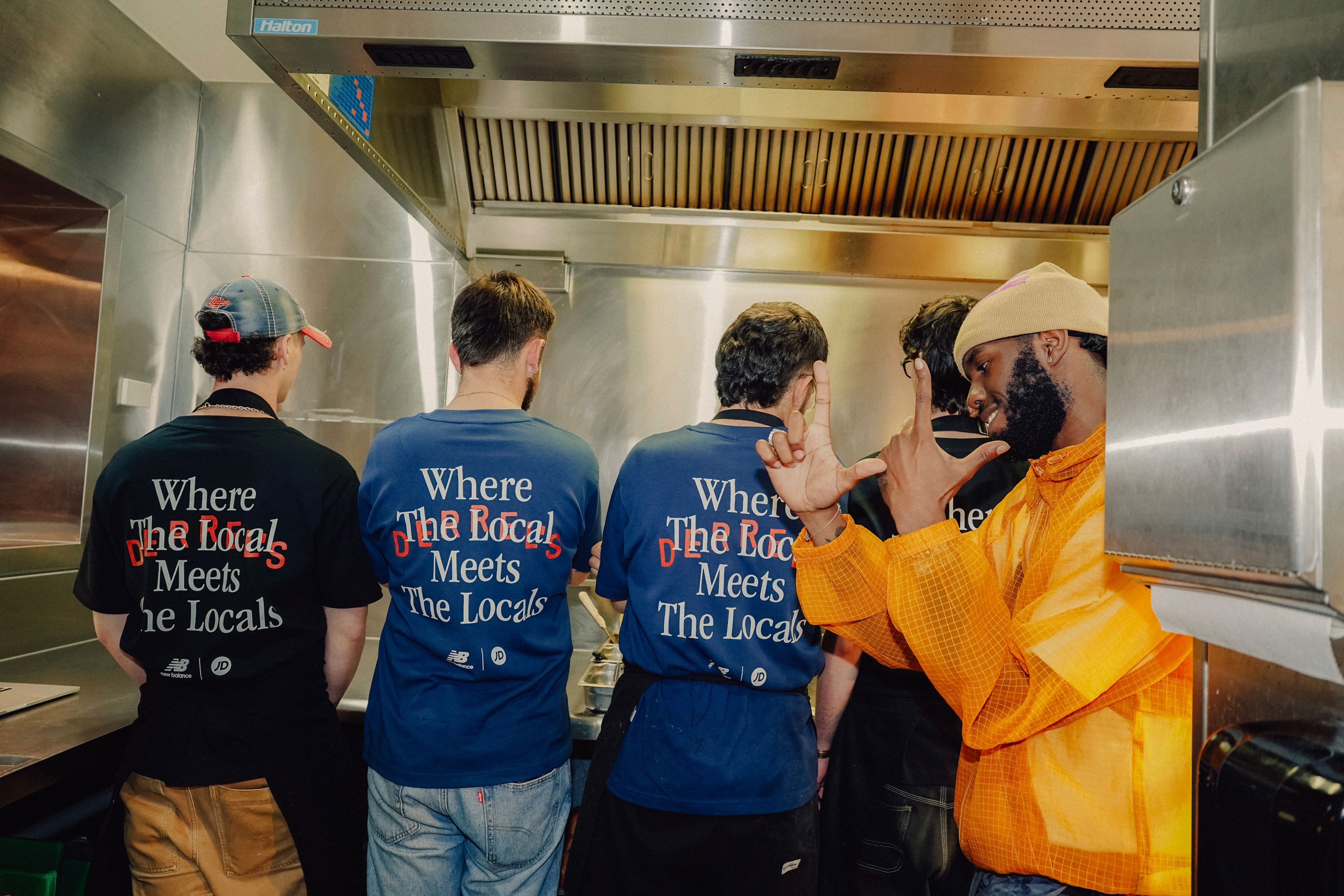 Three people from behind wearing blue denim jerseys printed with ‘Where the Local Meets the Locals’, facing Sainté in a yellow jacket