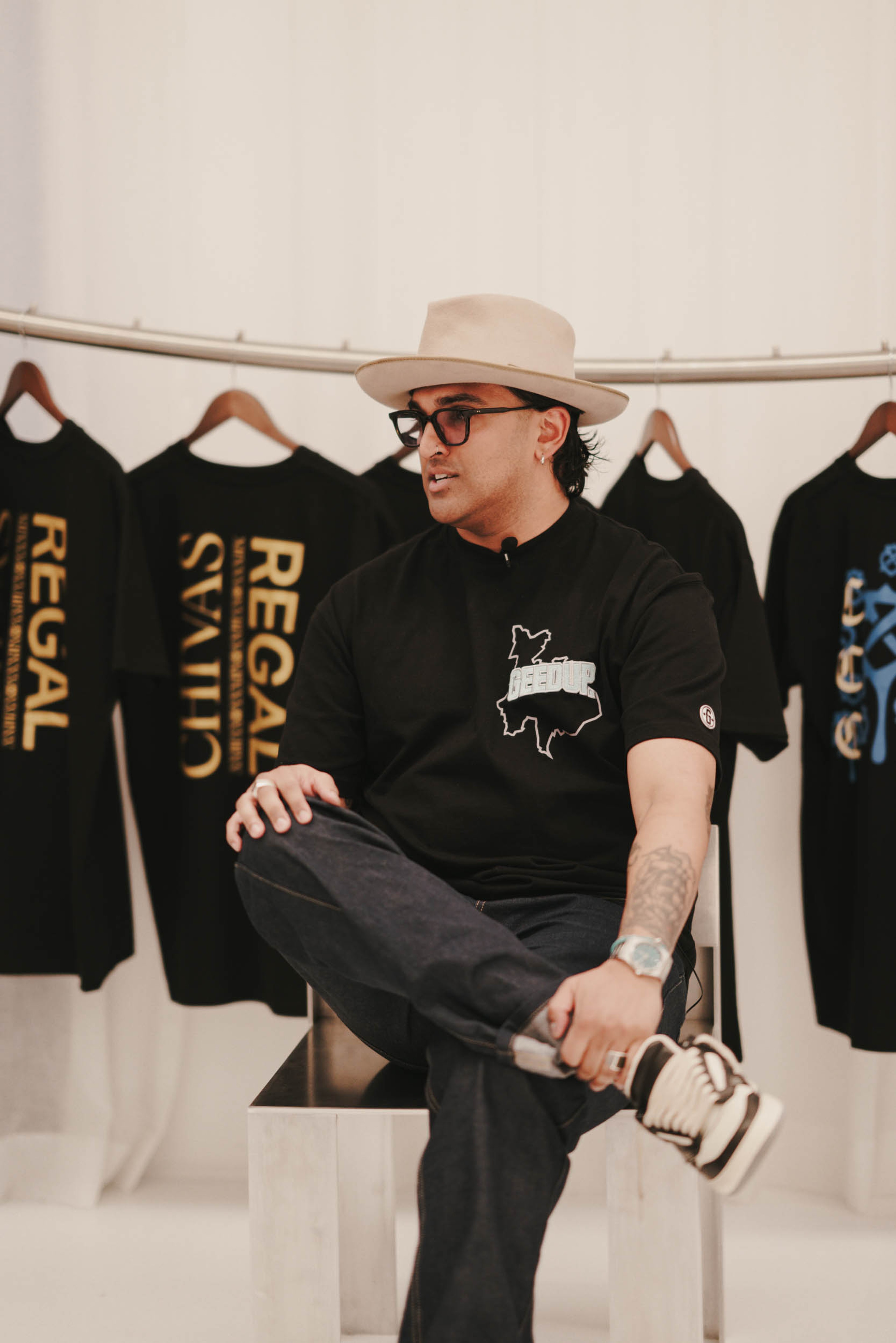 A man wearing a fedora, glasses, and a black graphic t-shirt sits in front of a rack of black t-shirts, some branded "Chivas Regal."