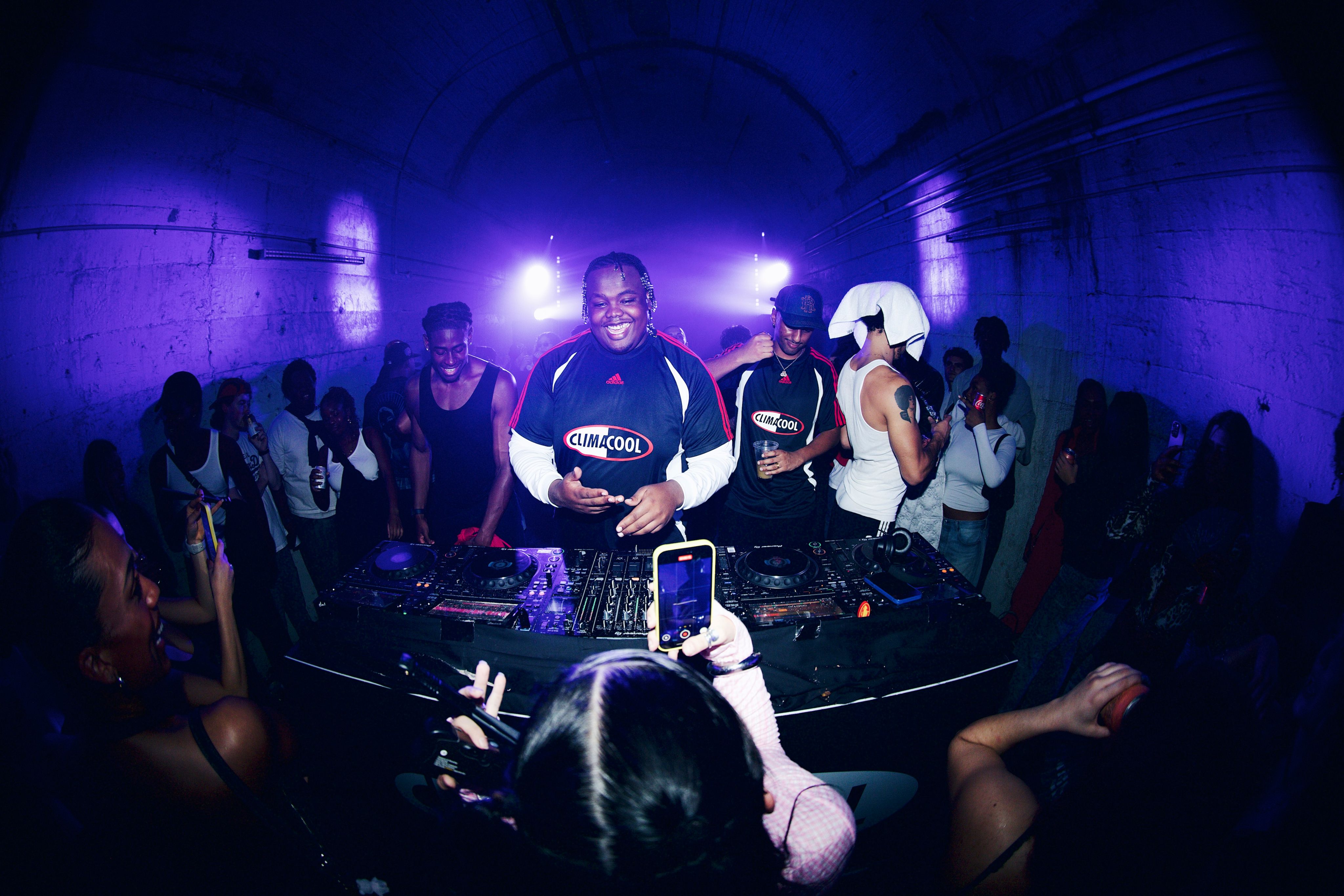 A DJ in a CLIMACOOL-branded jersey performing inside the Wynyard Tunnels under purple stage lights, surrounded by a crowd filming on their phones