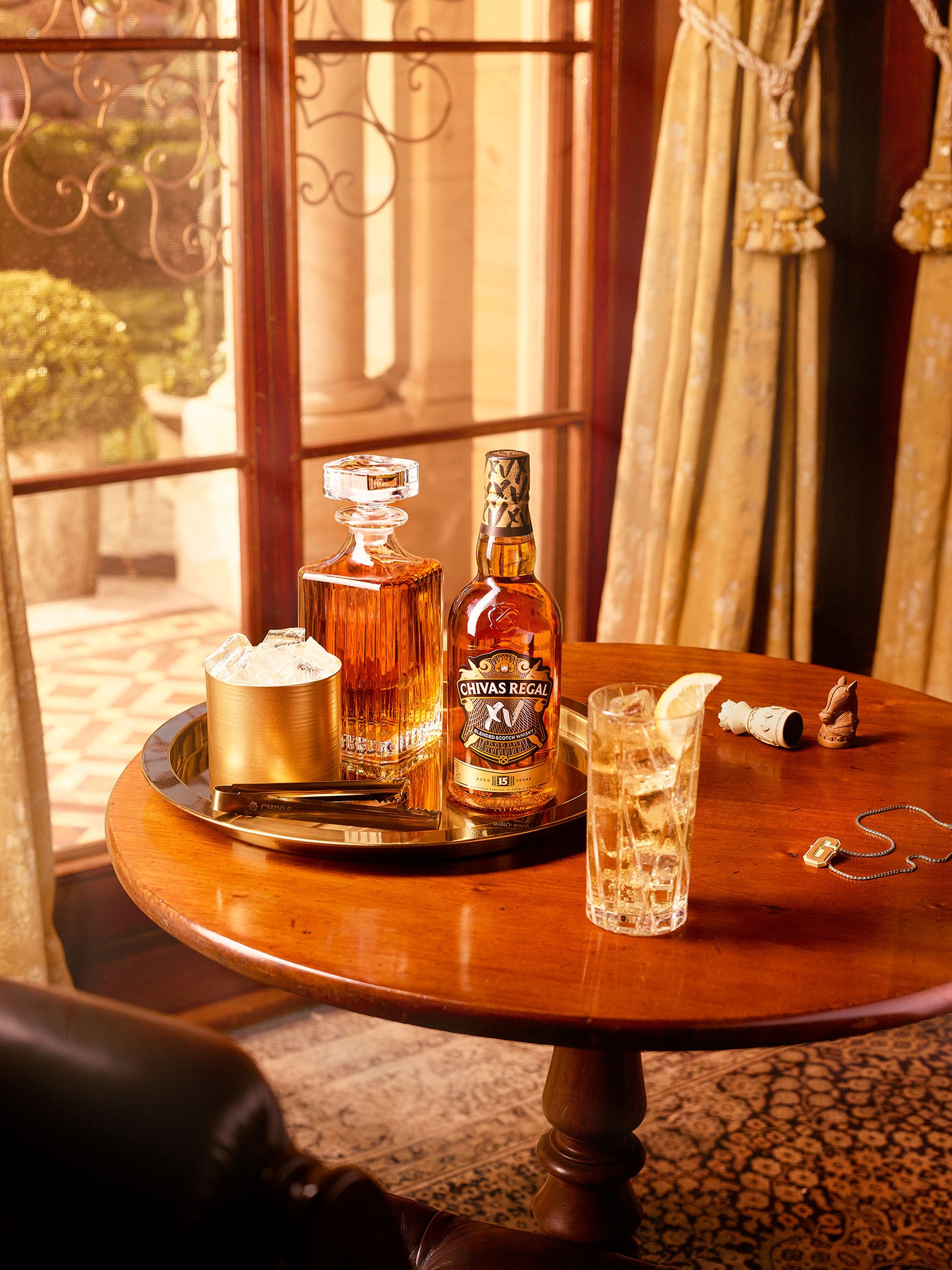A bottle of Chivas Regal XV, a whisky decanter, and an ice bucket on a golden tray, next to a tall glass of whisky with lemon, on a wooden table.