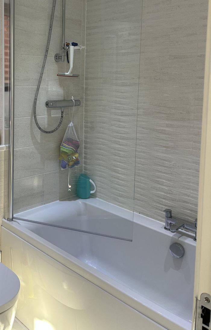 A bath with a hinged glass shower screen, thermostatic bar shower and textured wall tiles in a family bathroom.