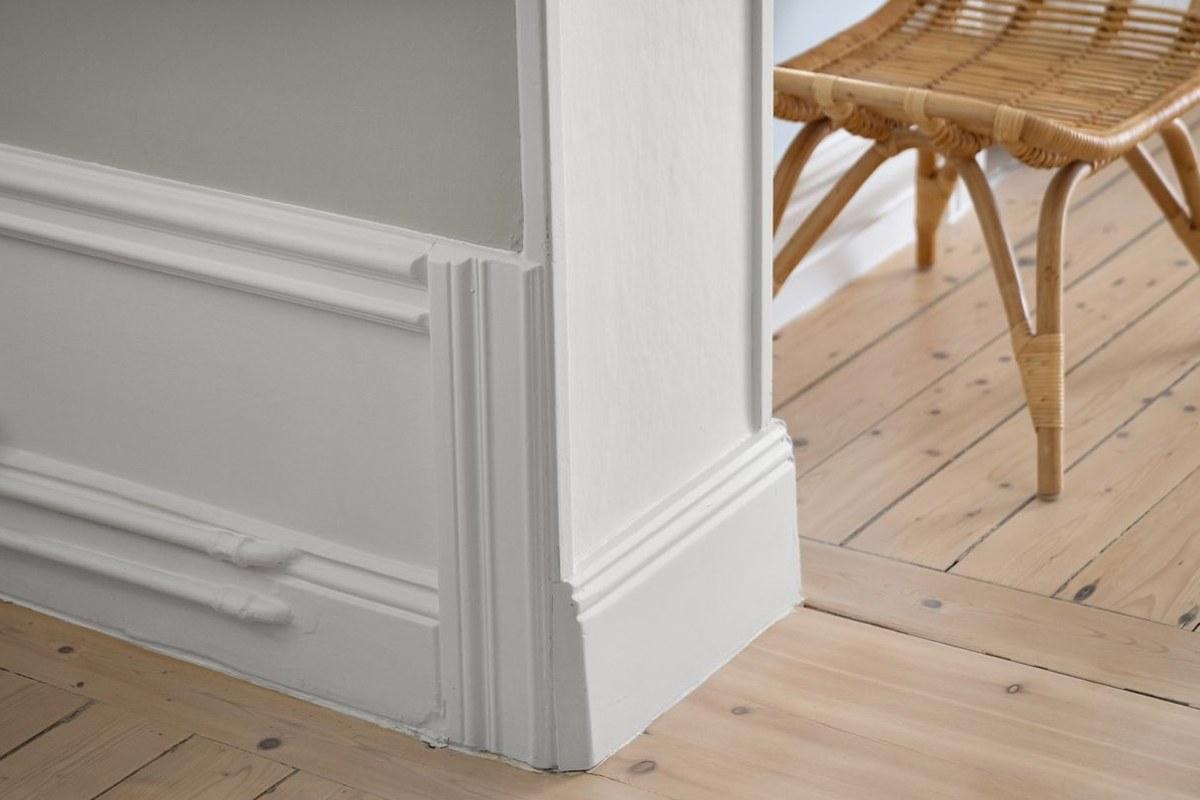 A tall white skirting board wrapping around a wall corner, with panelled moulding above pine floorboards and a rattan stool beside it.