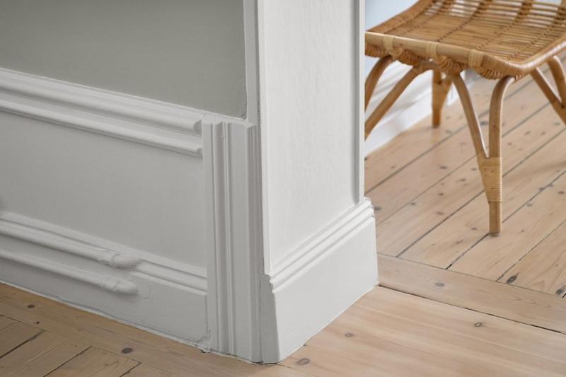 A tall white skirting board wrapping around a wall corner, with panelled moulding above pine floorboards and a rattan stool beside it.