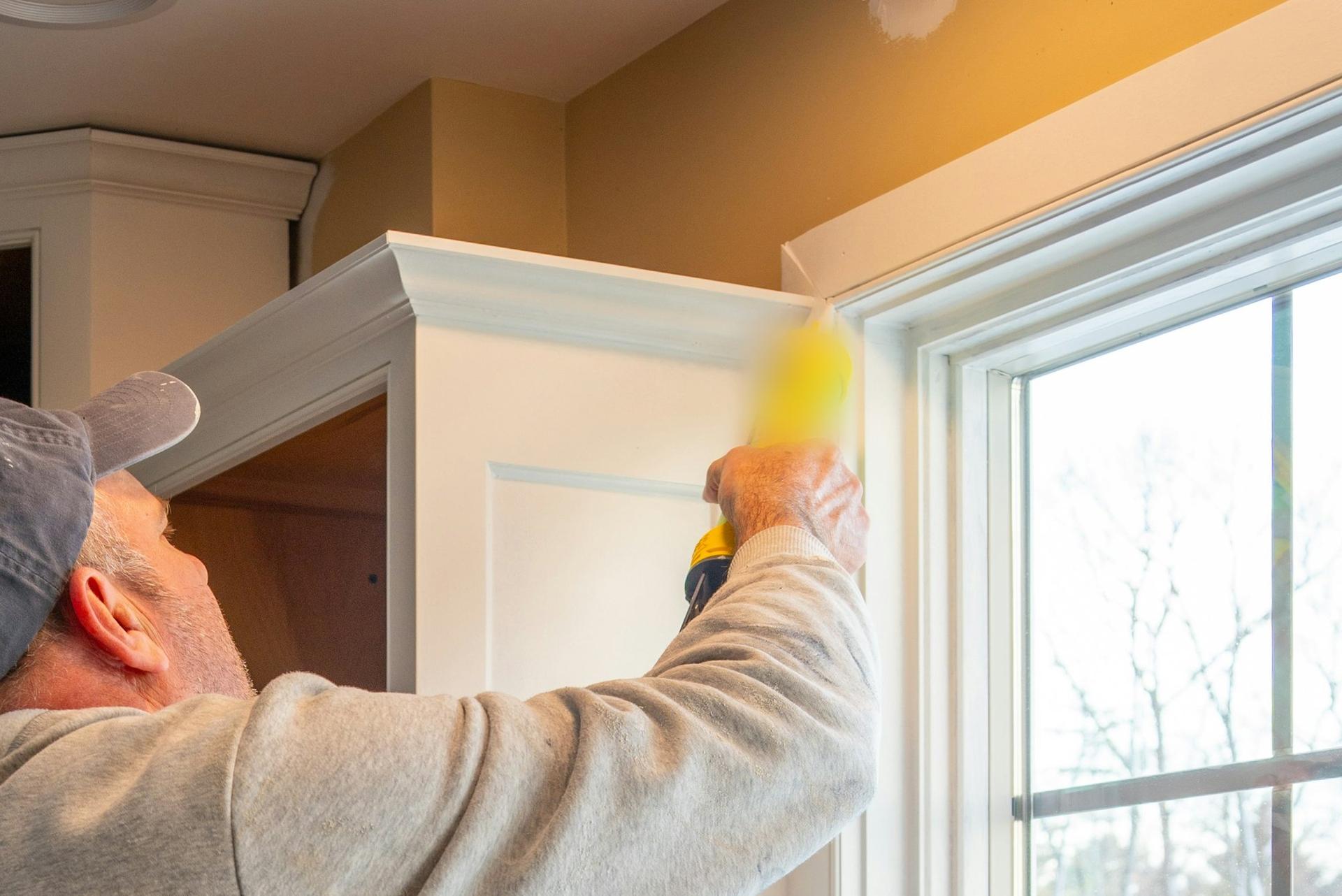 A tradesman in a cap and grey hoodie running a bead of caulk along the joint between a kitchen cabinet and a window frame.