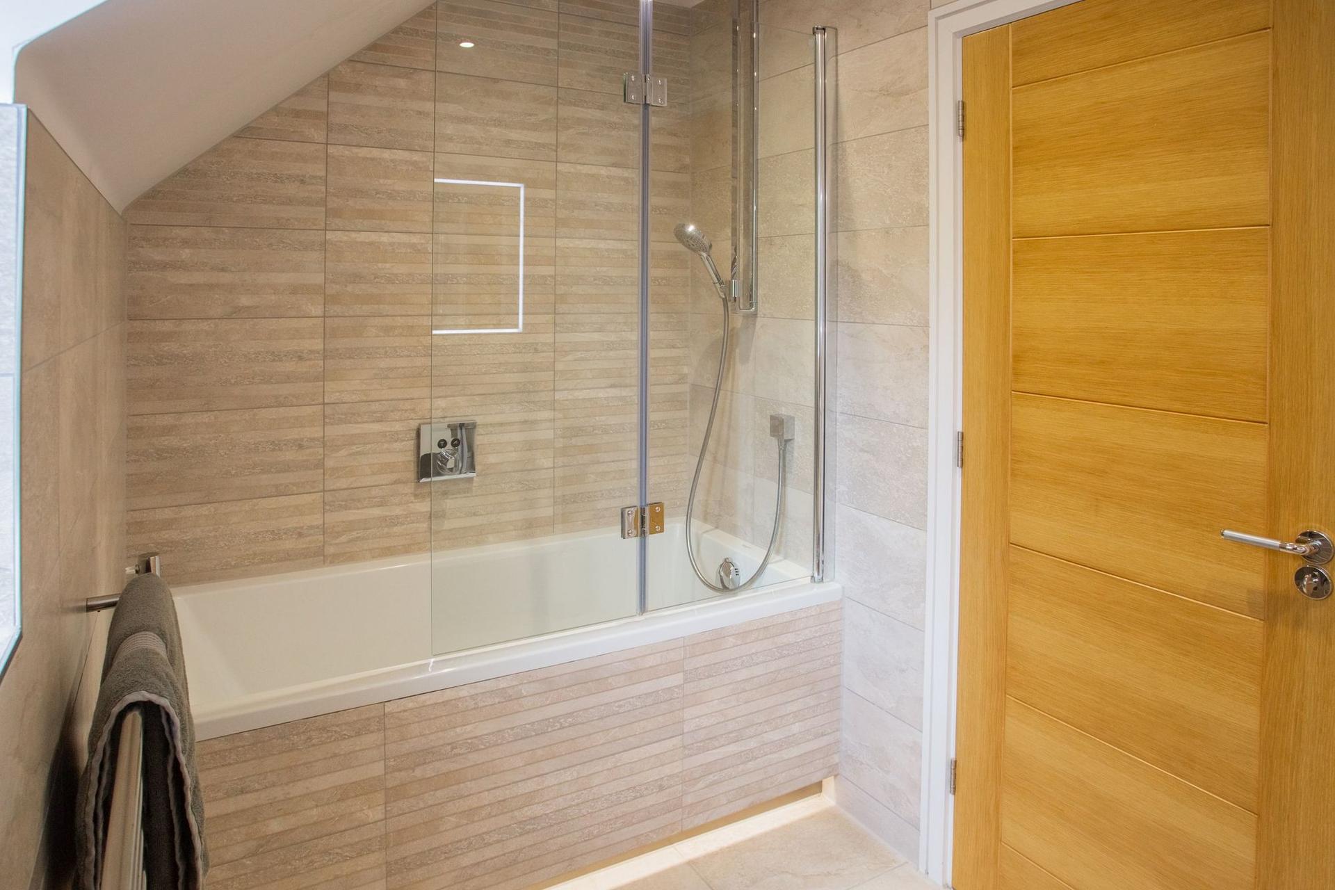 A finished bathroom with a tiled bath panel, folding glass shower screen, beige stone-effect wall tiles and an oak door.