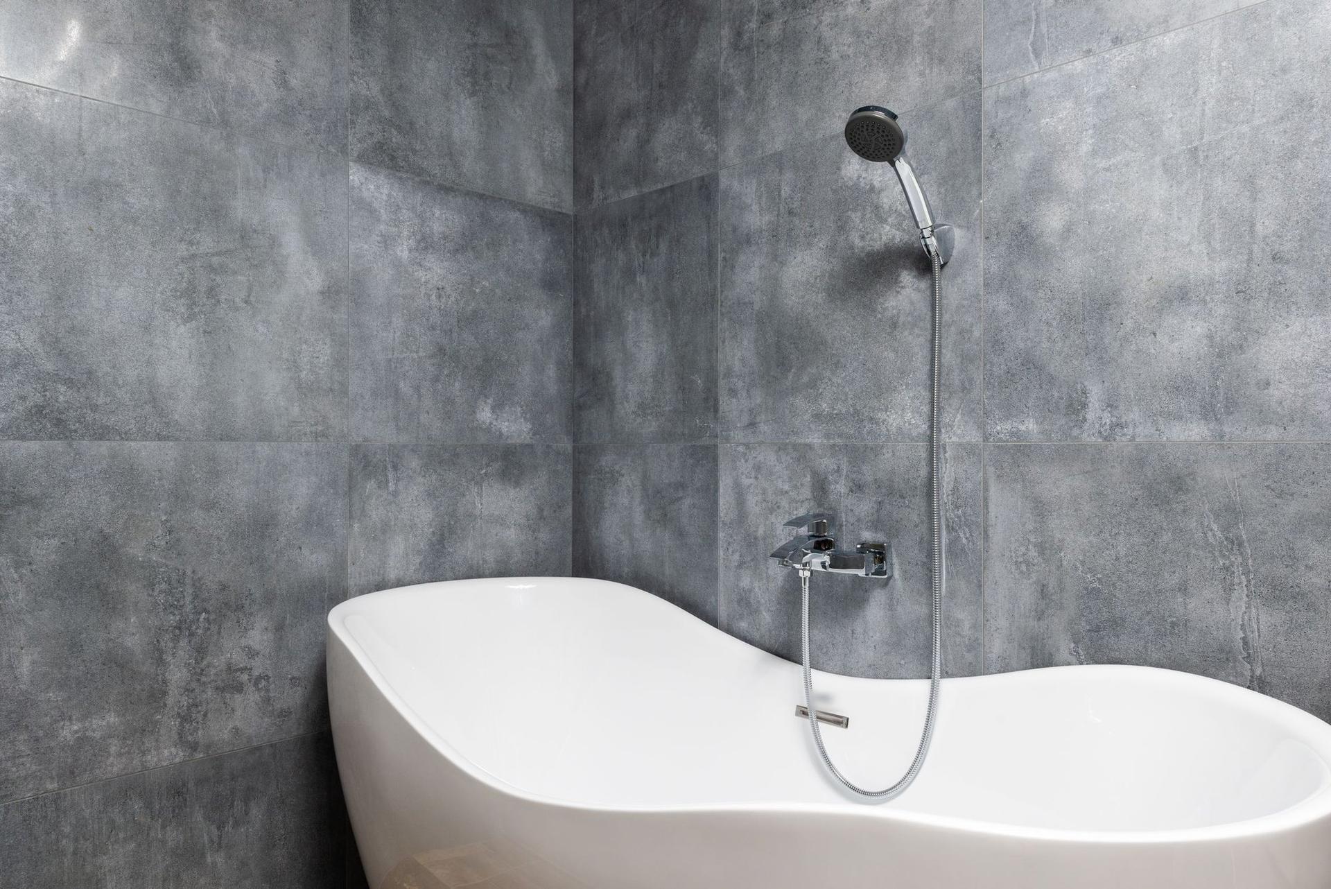 A white freestanding bath against large grey stone-effect wall tiles with a chrome wall-mounted mixer and hand shower.
