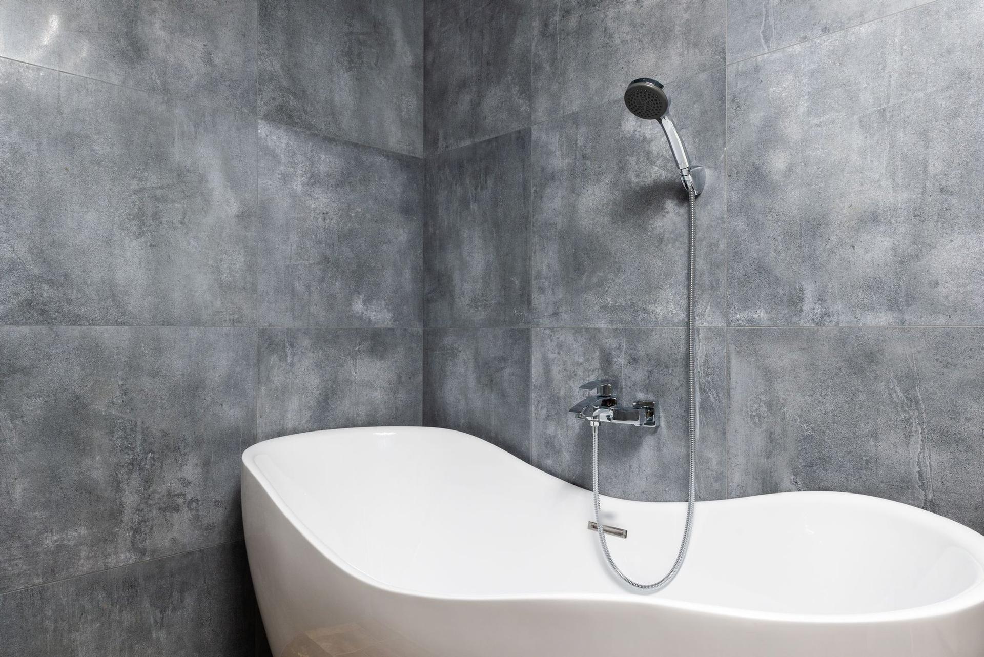 A white freestanding bath against large grey stone-effect wall tiles with a chrome wall-mounted mixer and hand shower.