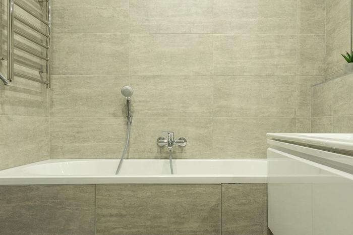 A white built-in bath with a tiled bath panel against large grey stone-effect wall tiles, with a chrome mixer and hand shower.