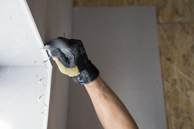A gloved hand drawing a blade along an internal wall corner, lifting away a thin strip of old material.
