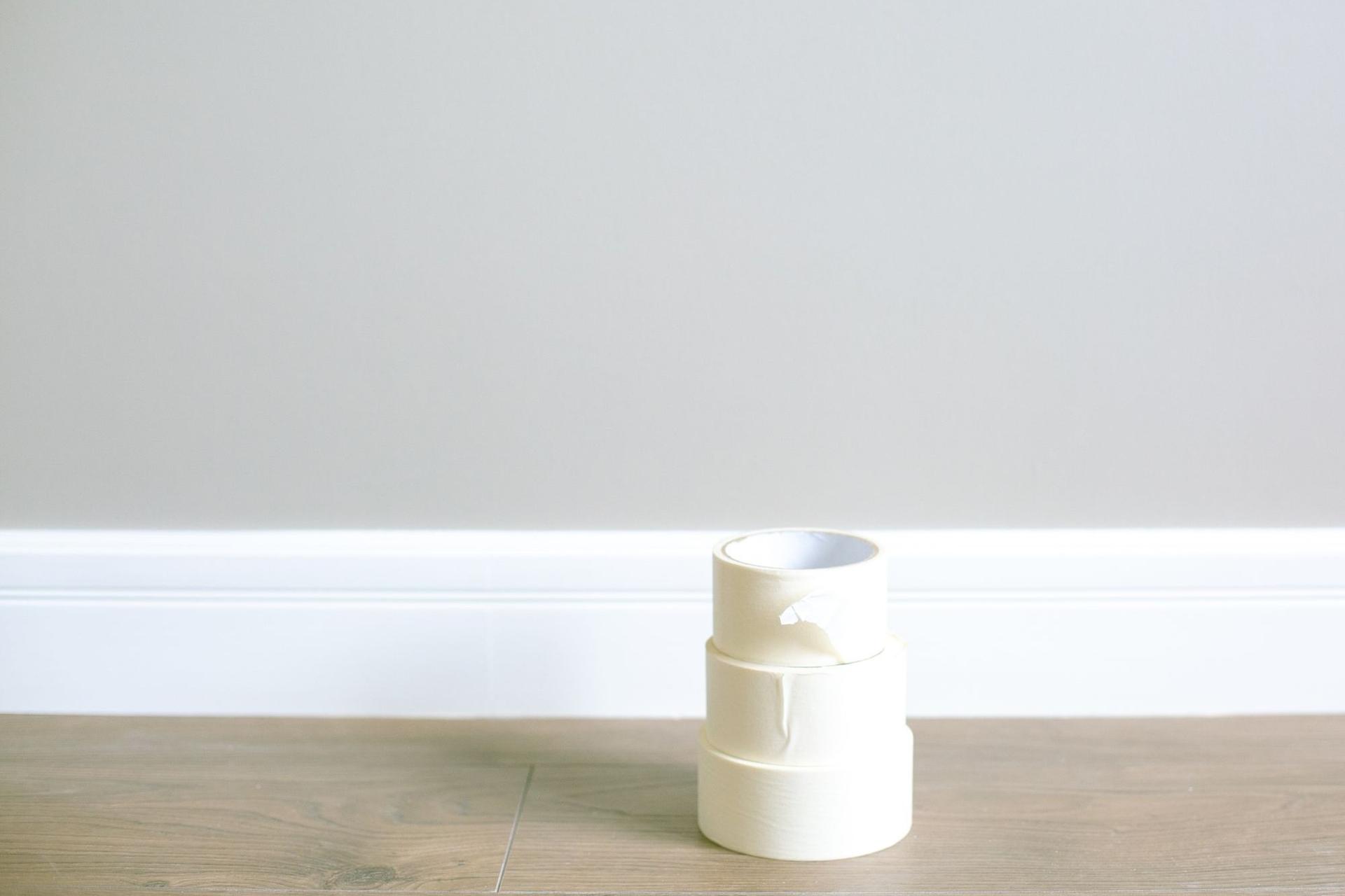 Three rolls of masking tape stacked on a wooden floor against a white skirting board and a plain grey wall.
