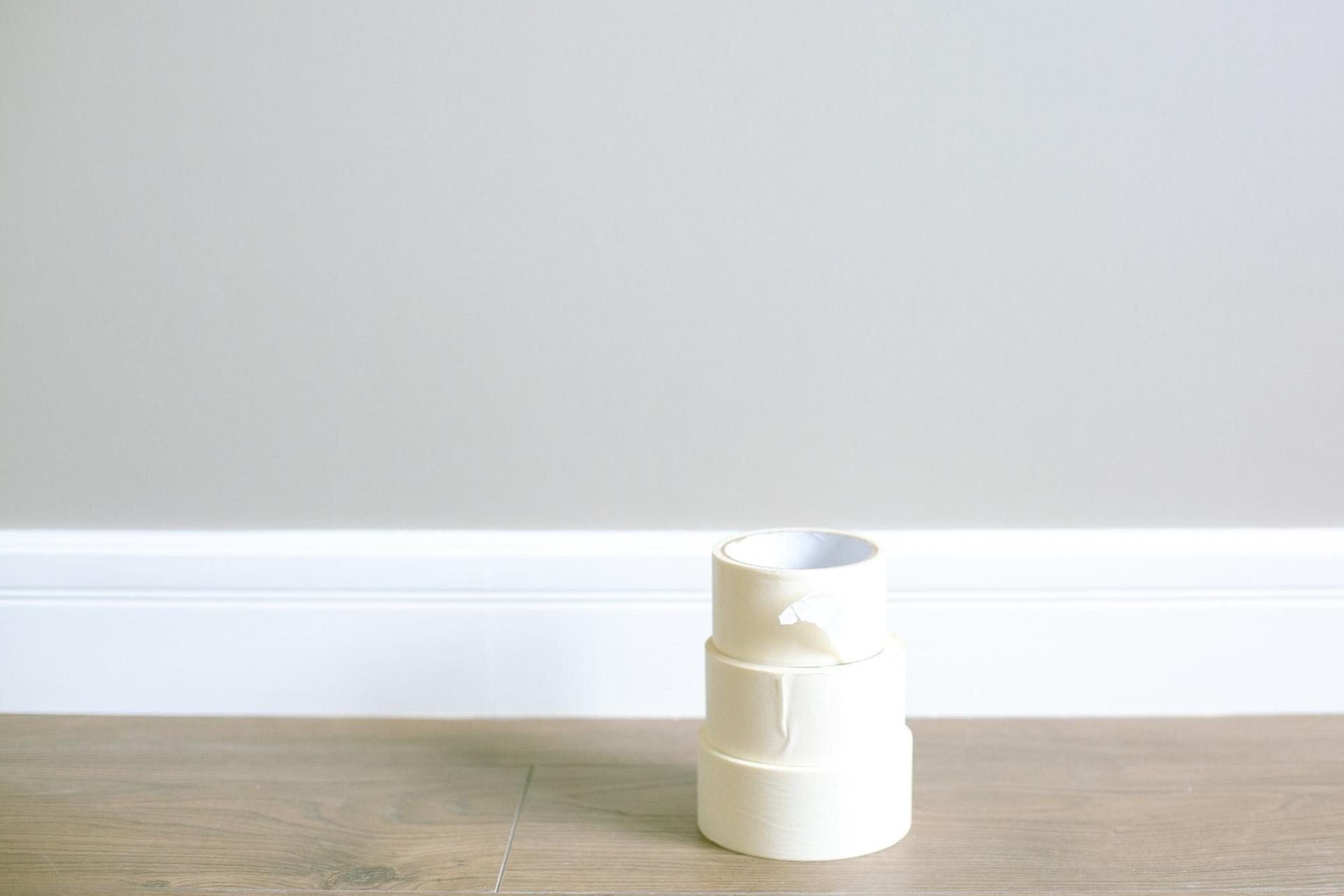 Three rolls of masking tape stacked on a wooden floor against a white skirting board and a plain grey wall.
