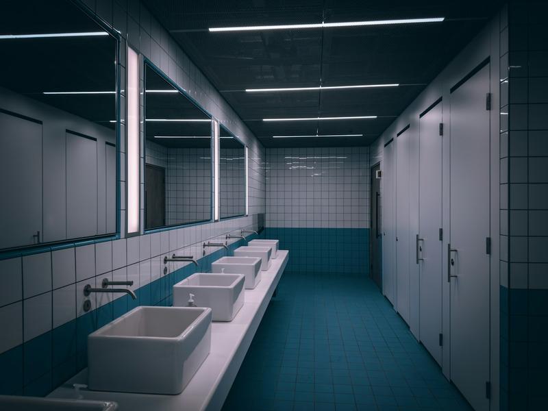 A row of square white basins with wall-mounted taps beneath long mirrors in a public washroom with teal floor tiles and white cubicle doors.