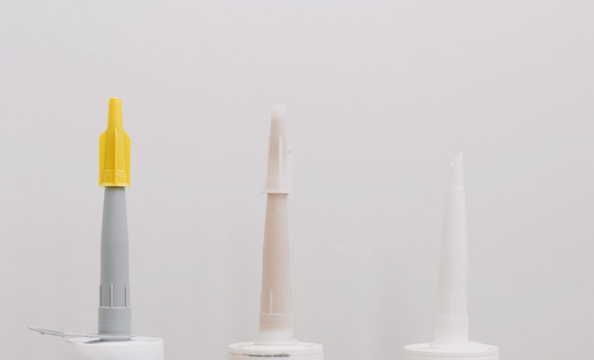 Three sealant cartridges with cut applicator nozzles lined up against a plain pale grey wall.