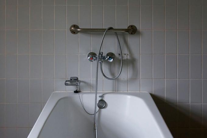 A white bath set against grey square wall tiles, with a chrome grab rail, mixer tap and hand shower.